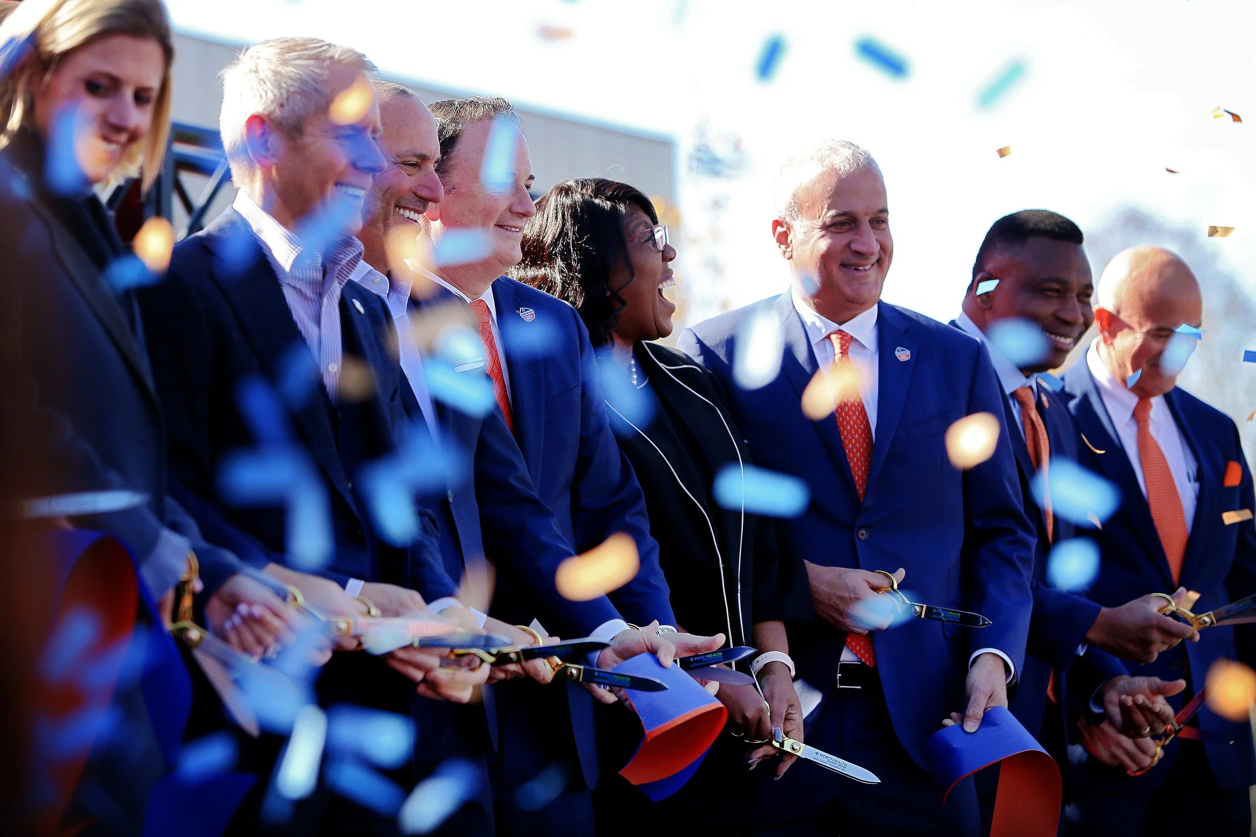 A group of well-dressed people participating in a ribbon-cutting ceremony, with confetti falling around them, outdoors under bright sunlight.