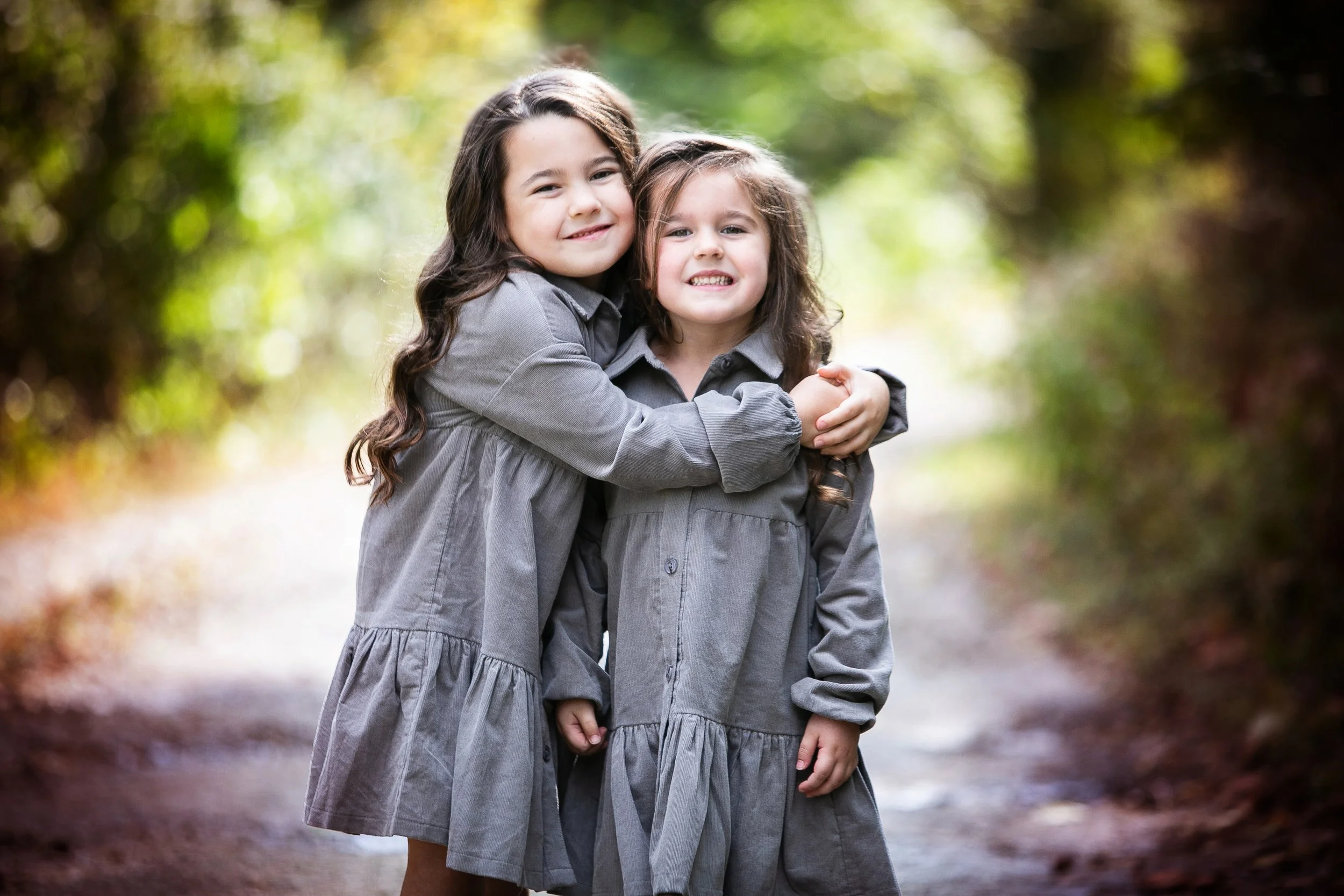 Two young girls hugging outdoors on a wooded trail, both wearing matching gray dresses, smiling at the camera.