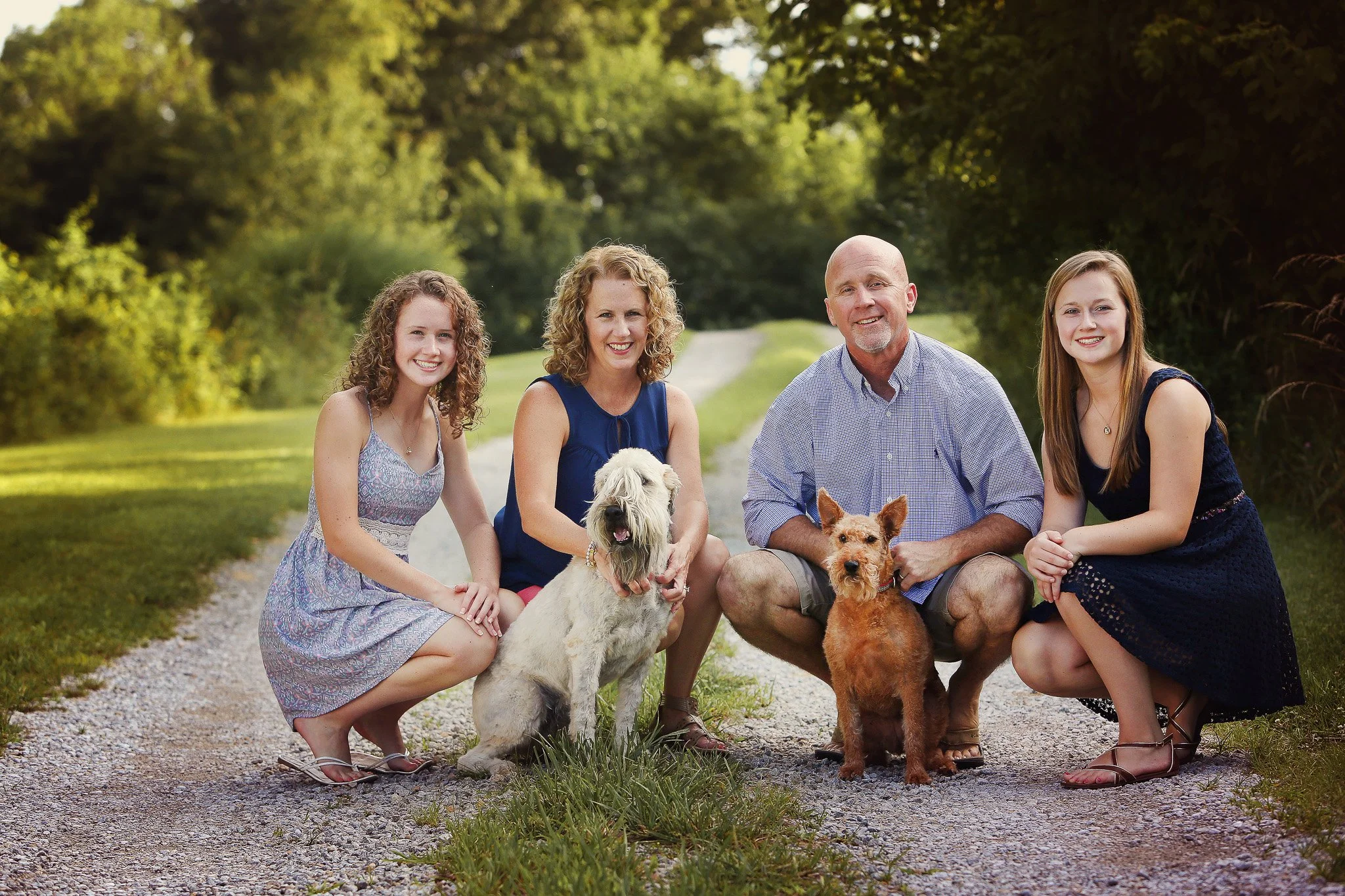 Family of five with two dogs outdoors on a gravel path surrounded by trees, smiling at the camera.