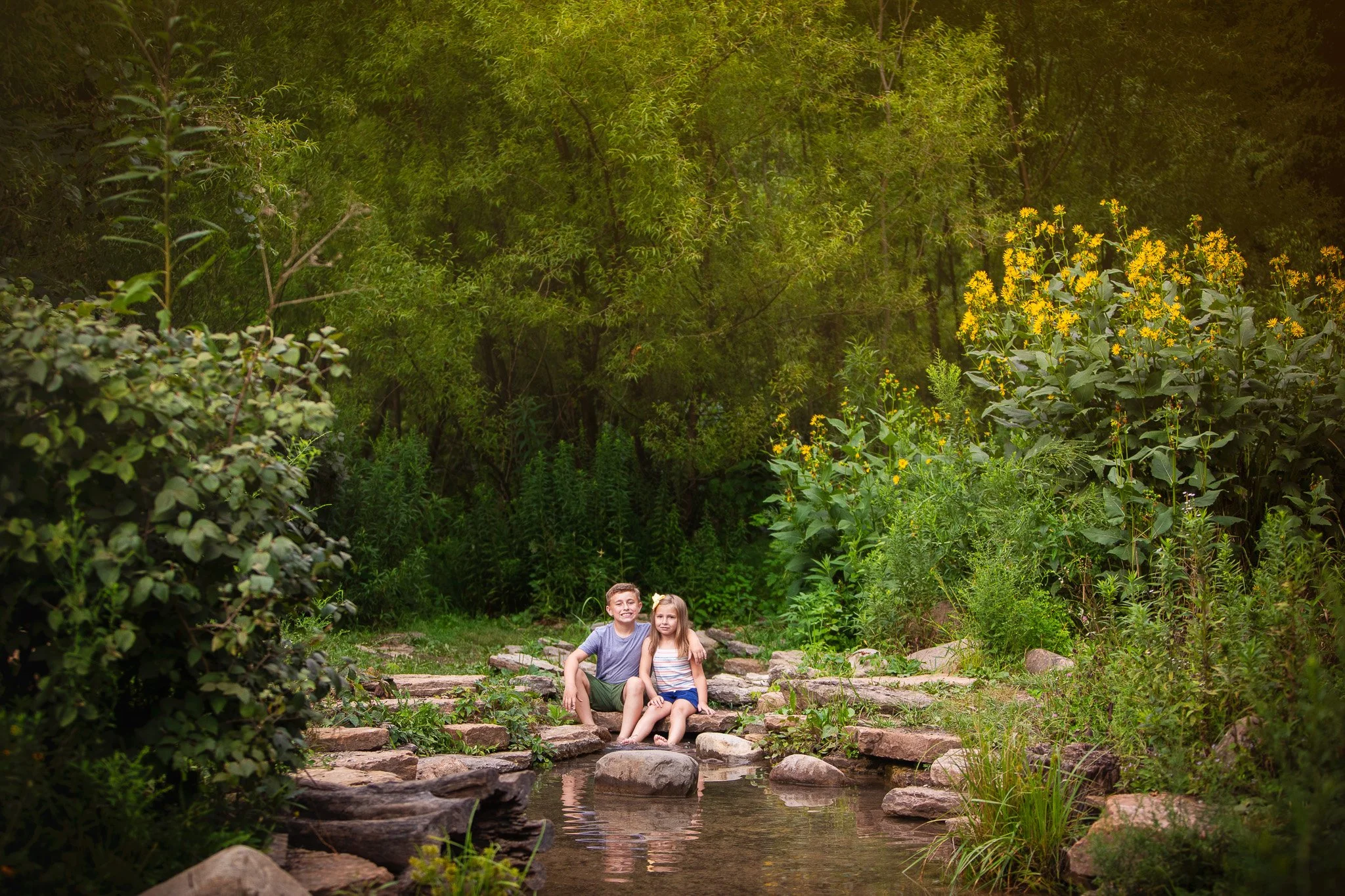 Two children, a boy and a girl, sit on rocks in a shallow creek surrounded by lush green bushes and tall trees, with yellow flowers on the right side.