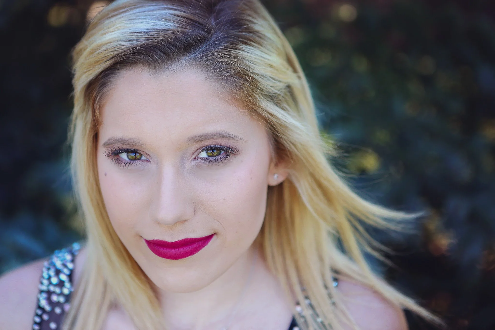 A close-up portrait of a woman with blonde hair, green eyes, and red lipstick, outdoors with a dark blurred background.