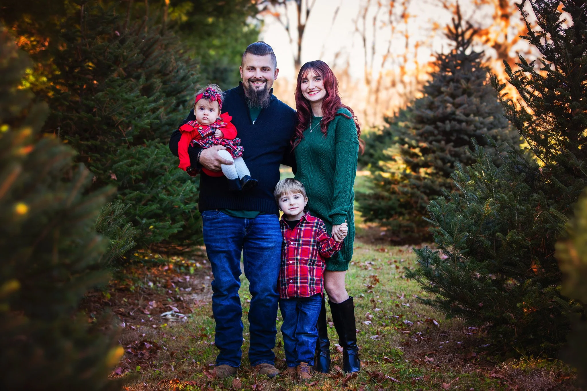 A family of four standing outdoors among Christmas trees, smiling at the camera during sunset. The father is holding a baby girl dressed in red, the mother is wearing a green sweater, and their older son is holding her hand, all dressed in festive cl