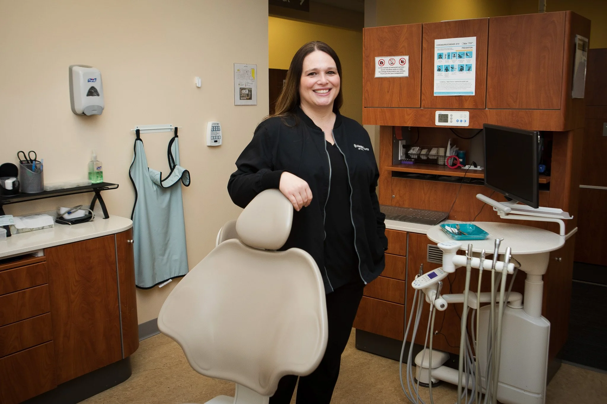 A female dentist or dental hygienist standing in a dental office, smiling, with dental equipment and furniture around her.