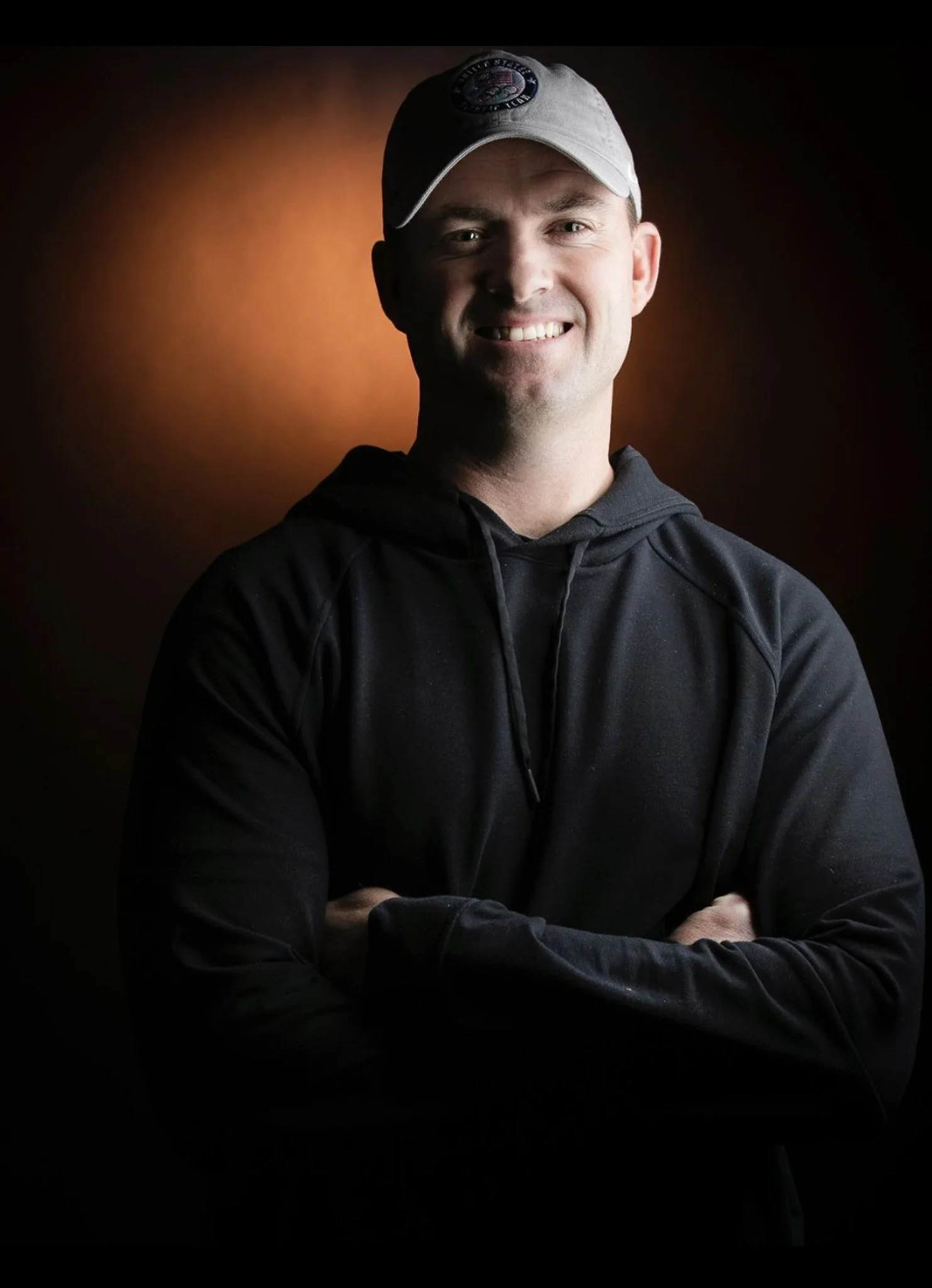 Portrait of a smiling man wearing a white baseball cap and black hoodie, standing with arms crossed against a dark background with a gradient orange glow.
