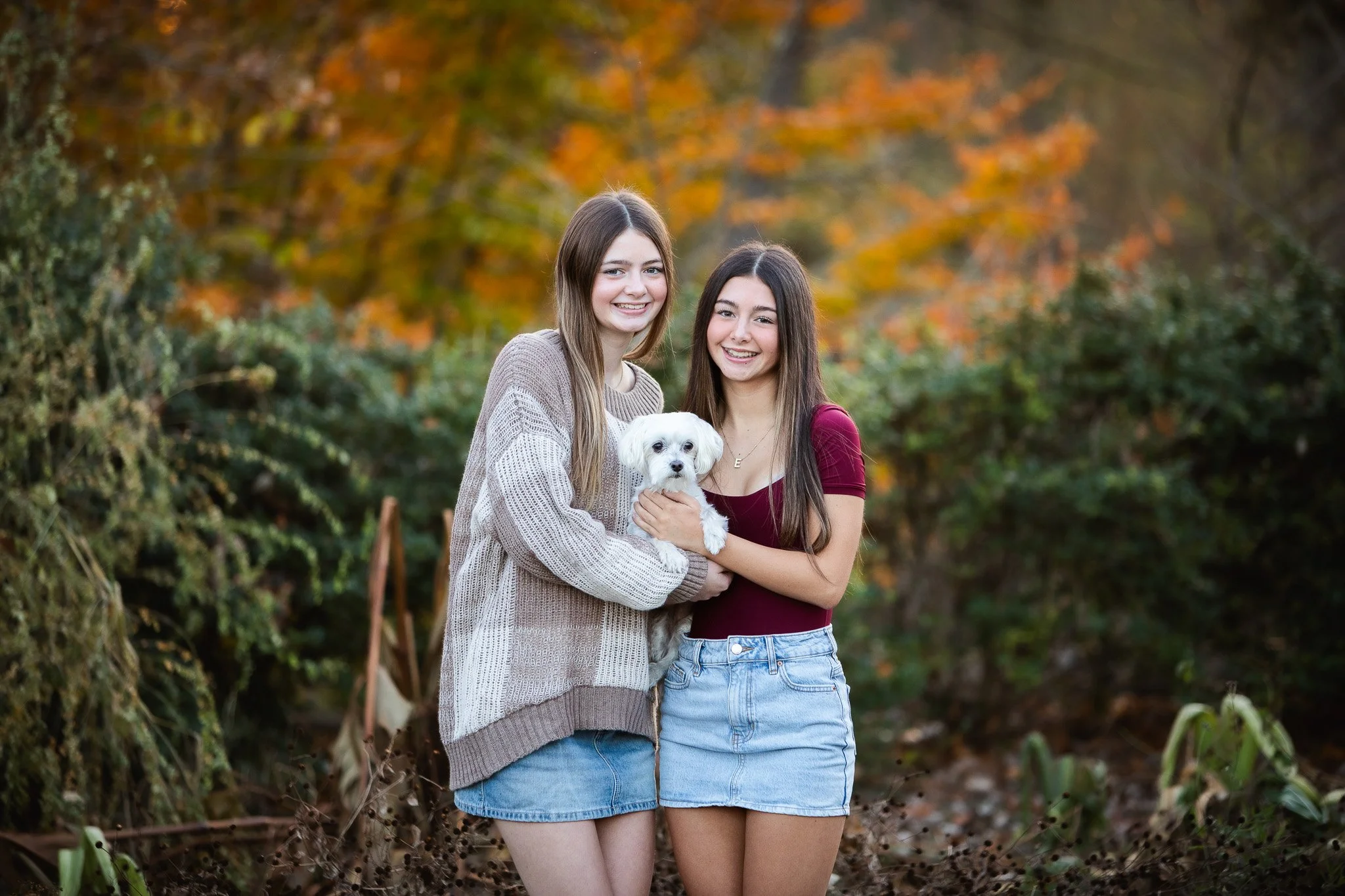 Two young women standing outdoors with autumn foliage in the background, smiling, holding a small white dog.