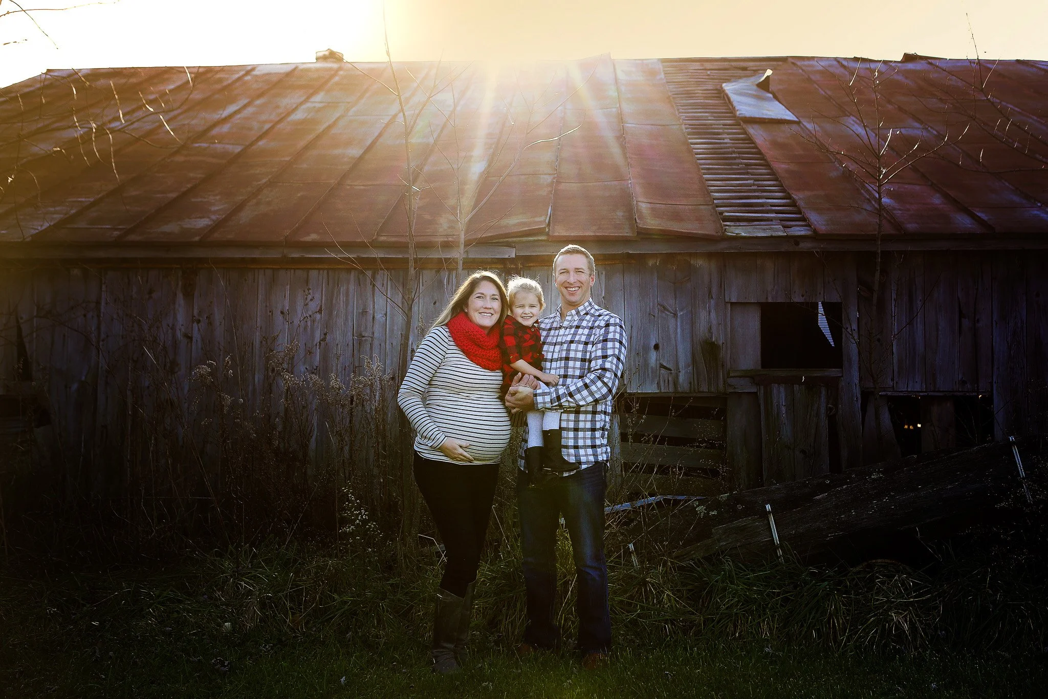 A family of three standing in front of an old barn during sunset, with a pregnant woman, a man, and a young girl smiling.