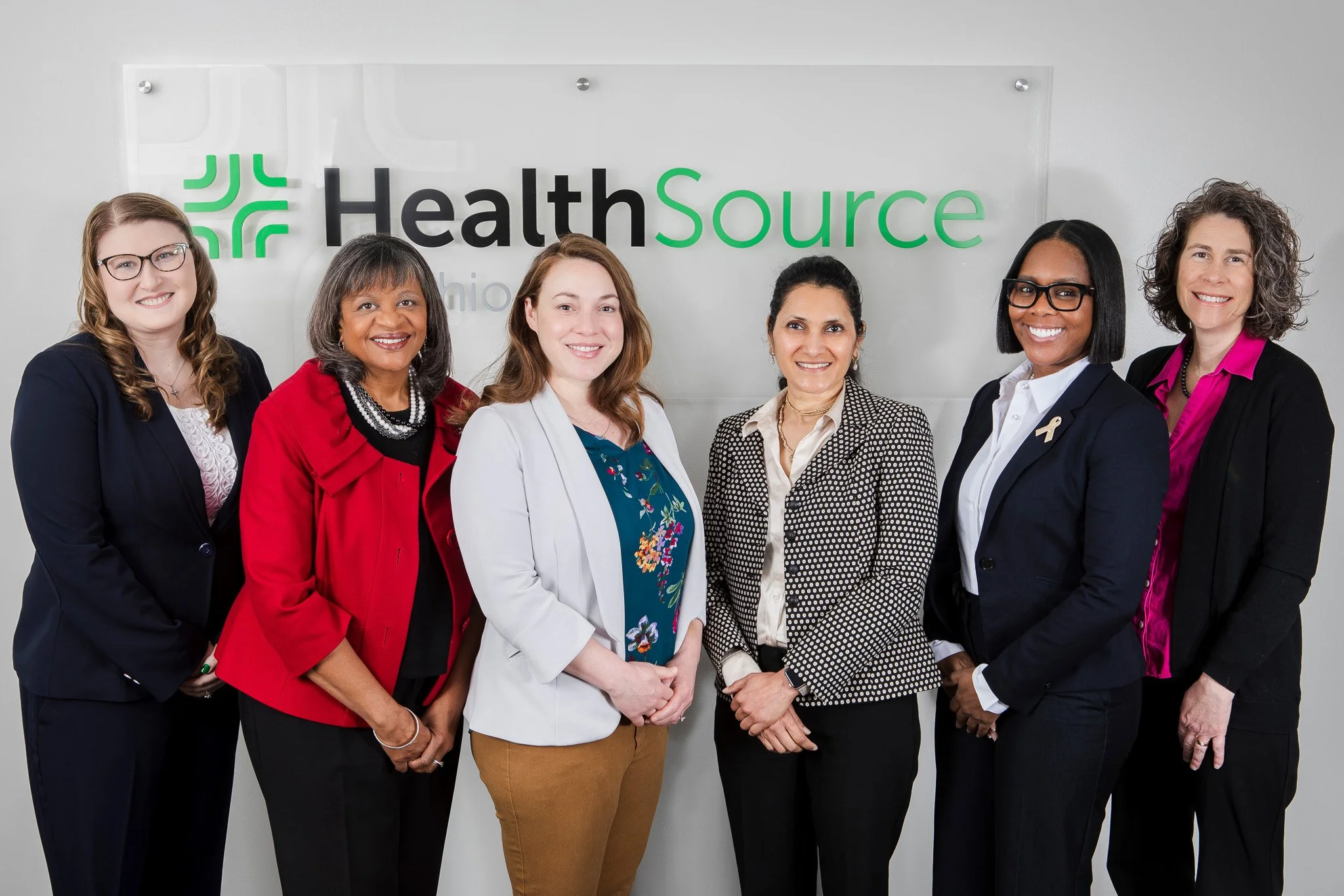 Group of six women standing in front of a 'HealthSource Ohio' sign, smiling, dressed in professional attire.