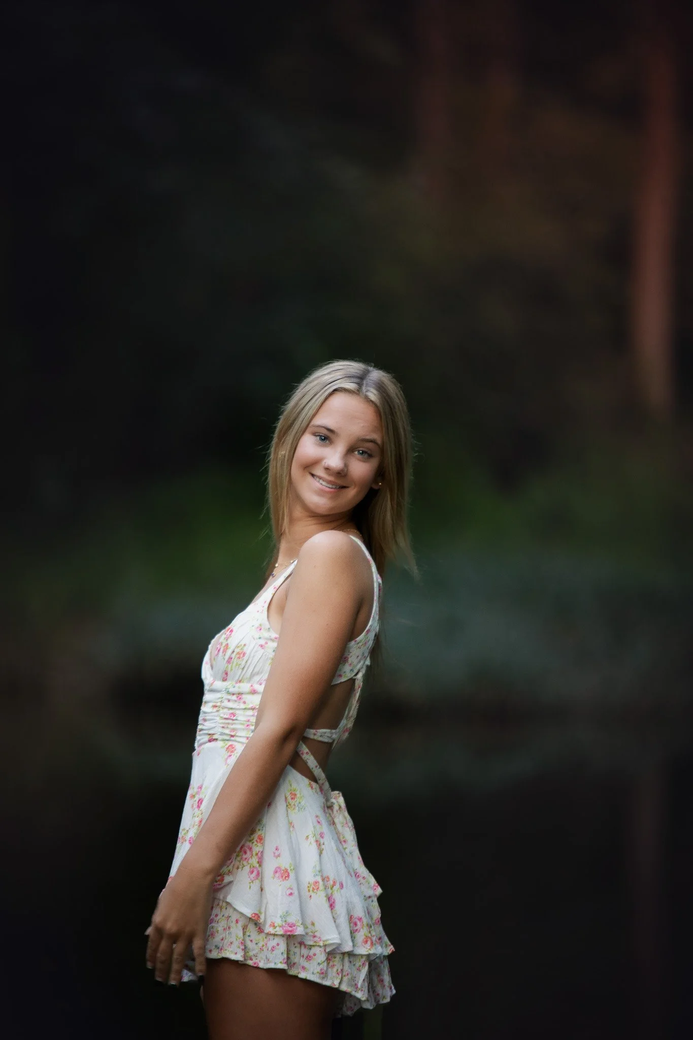 Young woman with blonde hair smiling outdoors at dusk, wearing a floral dress with cutouts.