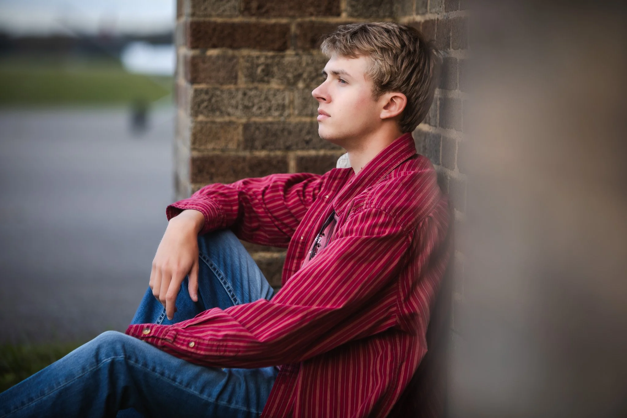 Young man in a red striped shirt sitting on the ground, leaning against a brick wall, looking into the distance with a contemplative expression.