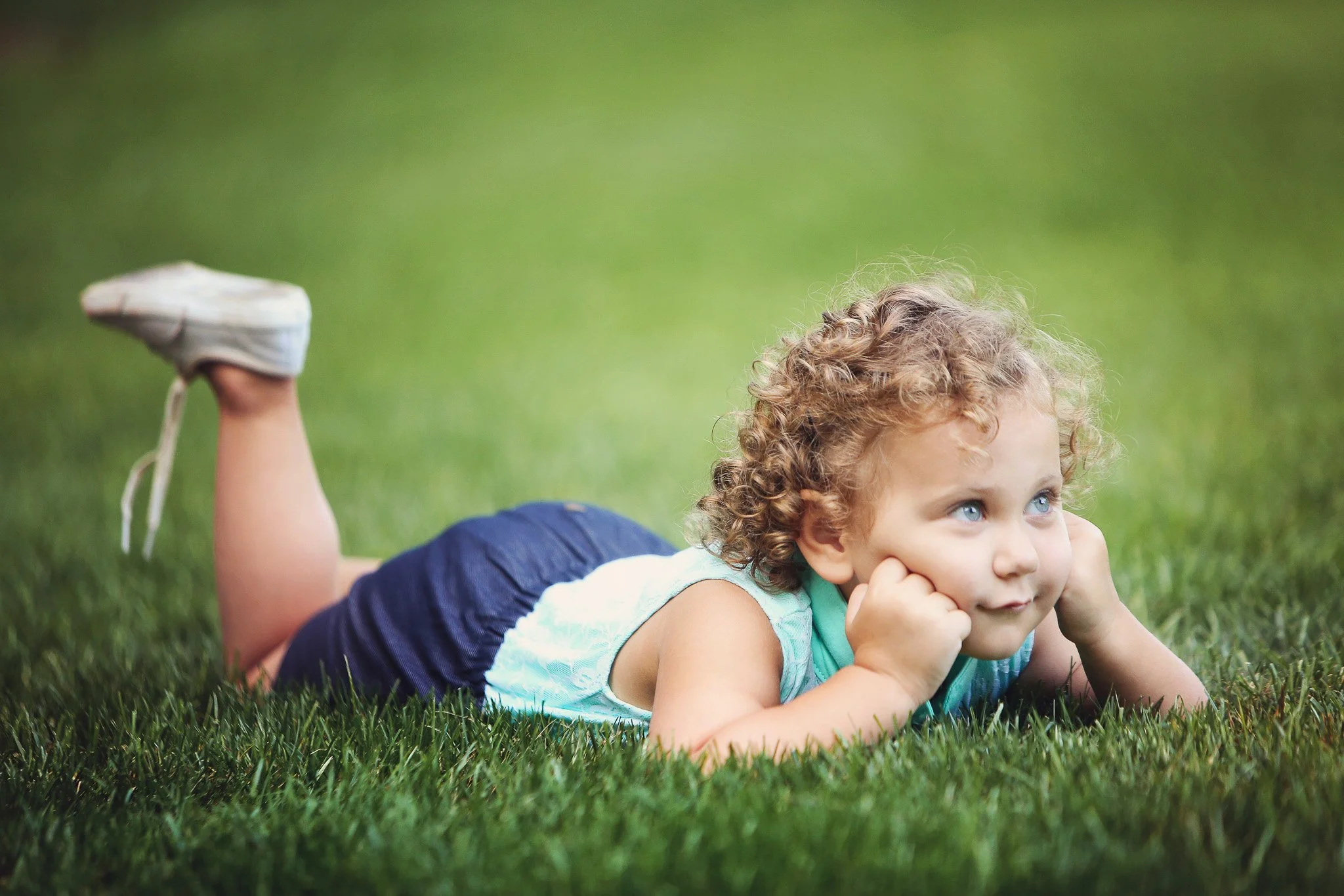 Young child with curly hair lying on grass with chin resting on hands, looking to the side, outdoors.