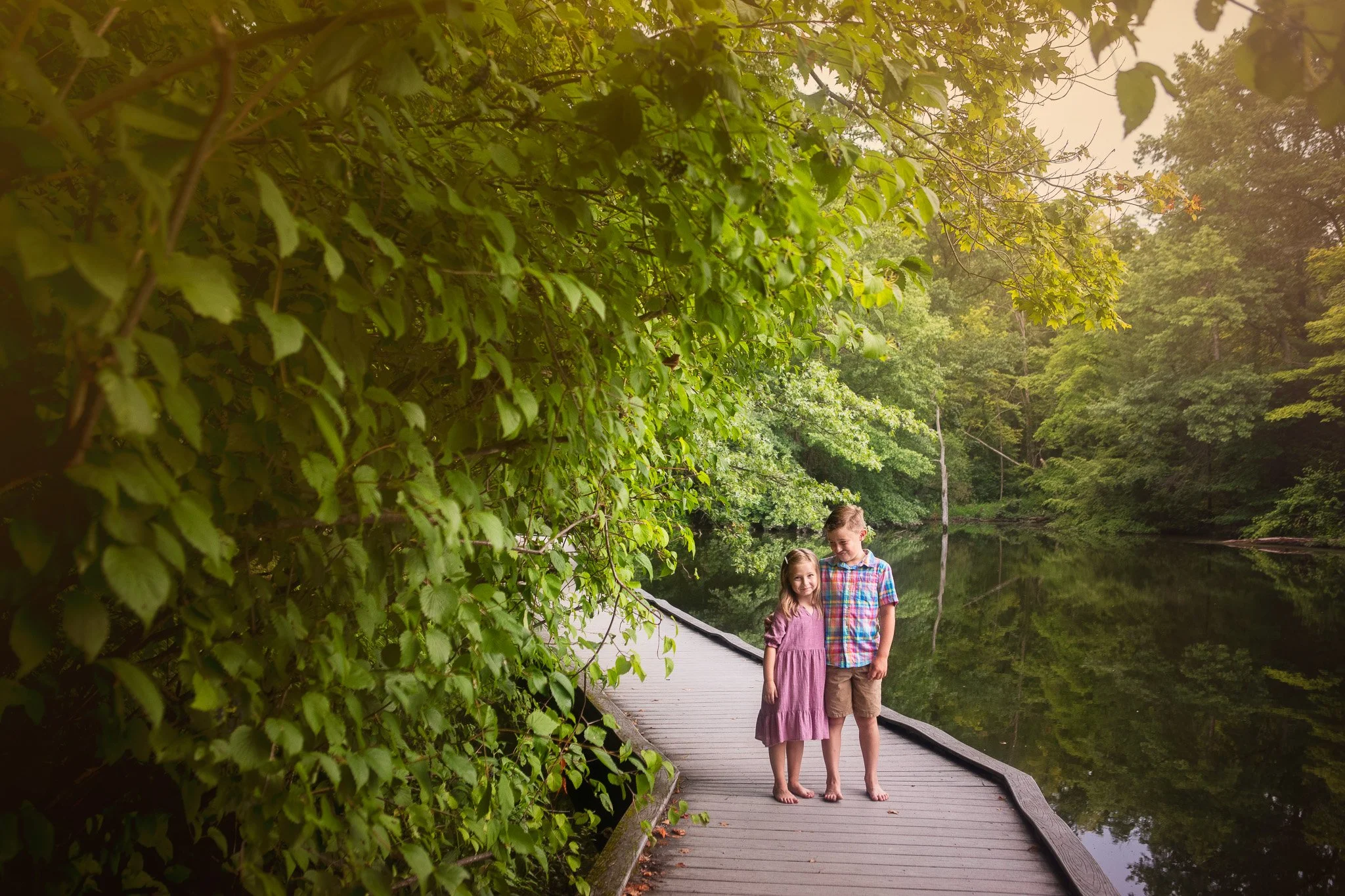 Two children standing on a wooden pathway beside a calm lake, surrounded by lush green trees and foliage.