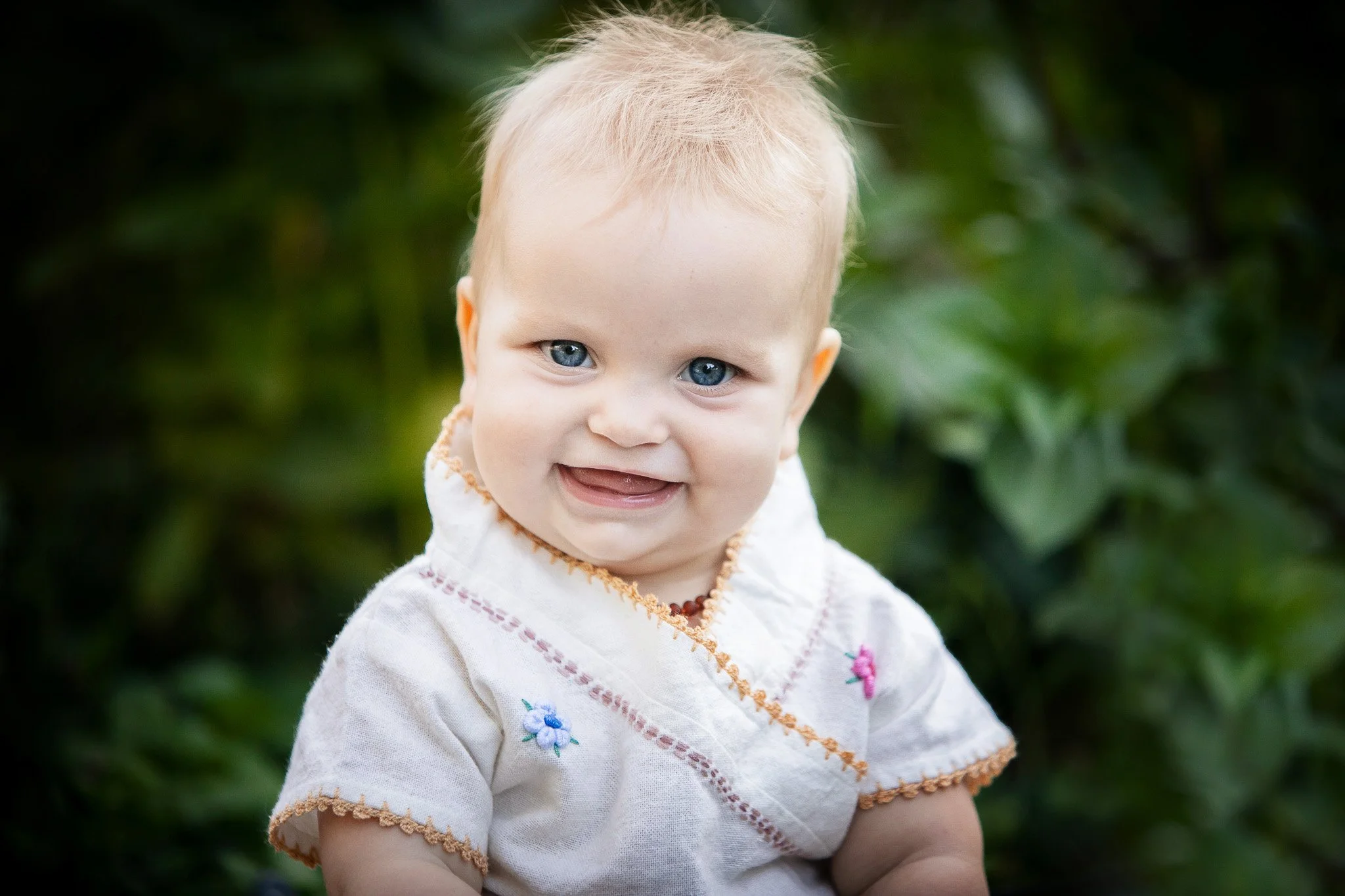 A smiling baby with light blonde hair and blue eyes, wearing a white embroidered shirt, outdoors with green foliage in the background.