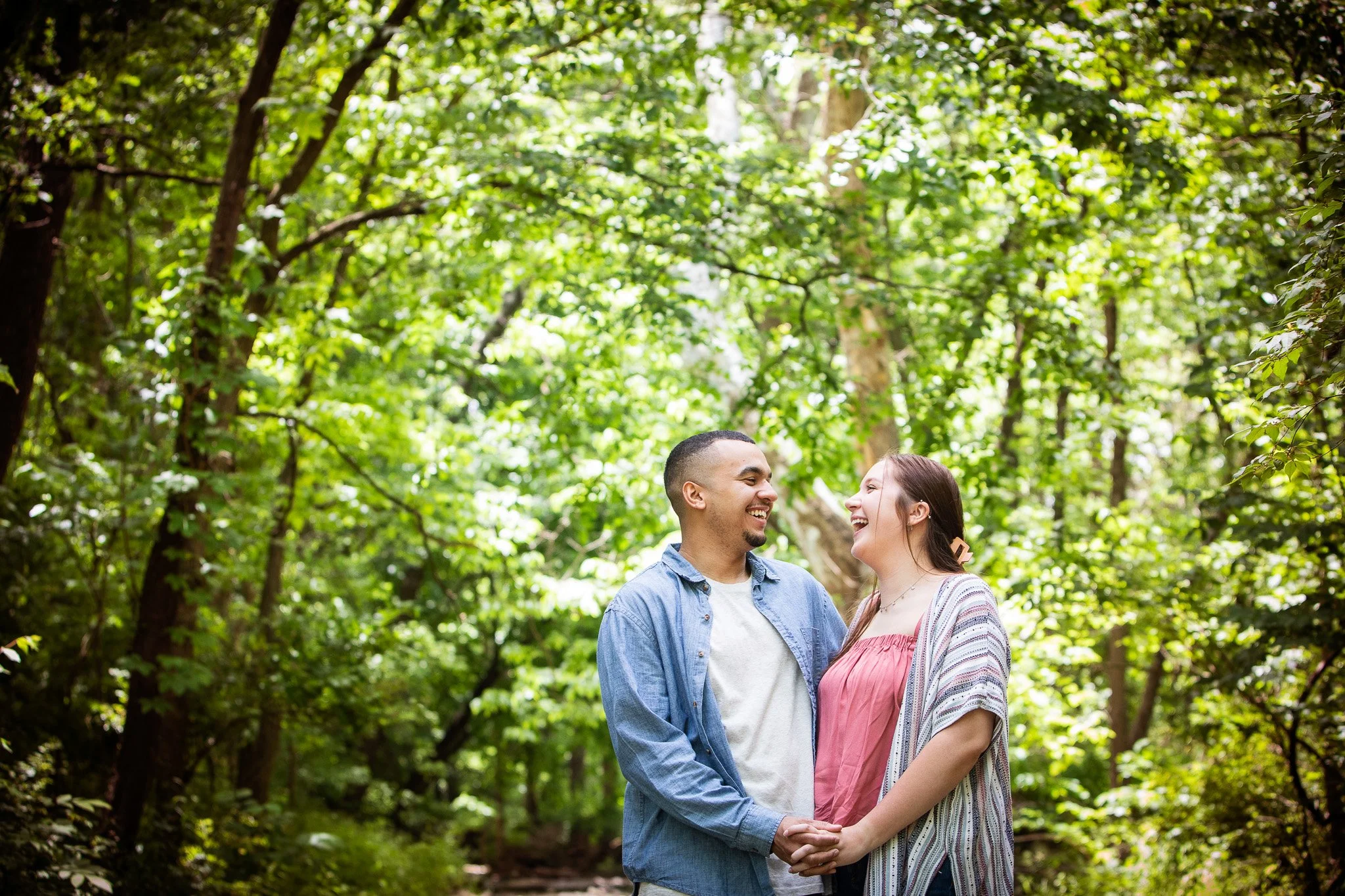 A couple holding hands and smiling at each other in a lush, green forest.