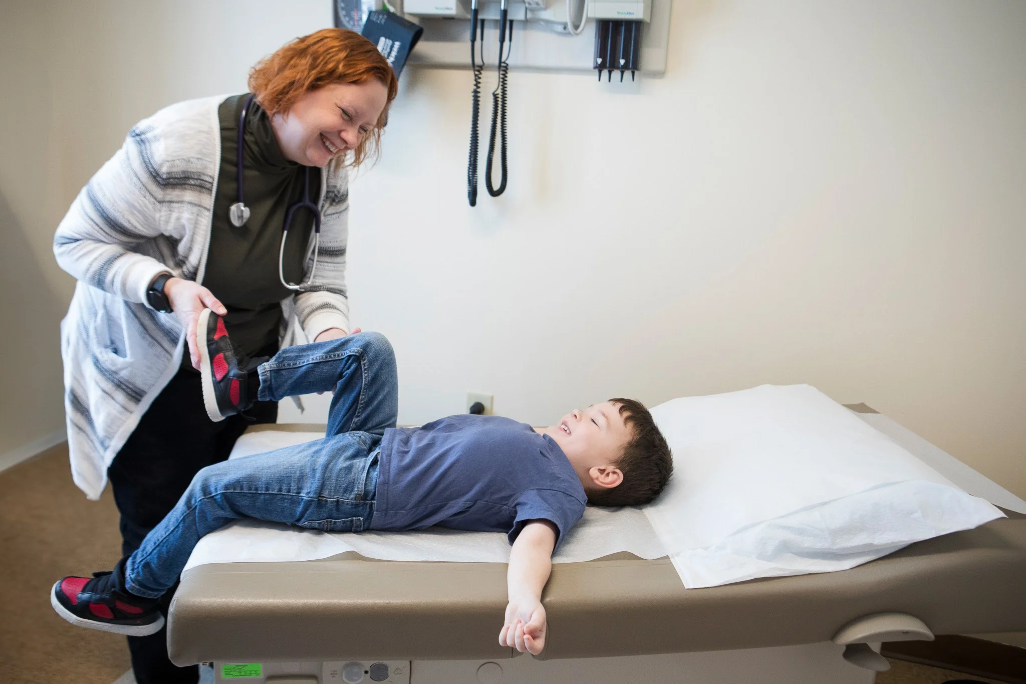 A female healthcare worker laughing while lifting a child's leg on an examination table in a medical office.