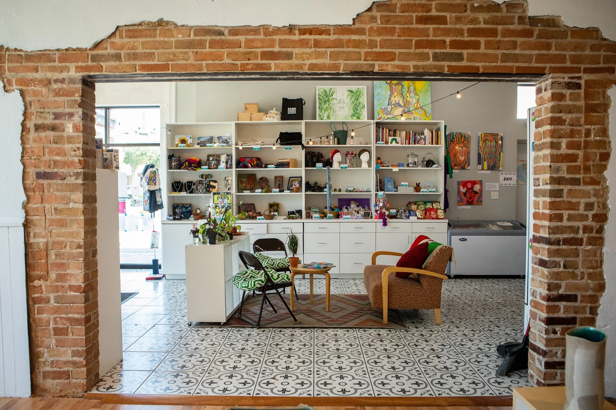 Interior view of a small shop or cafe with white shelves displaying various items like jewelry, art, and accessories. There is a seating area with a small table, a brown armchair with red cushion, chairs with green and white patterned covers, and pot