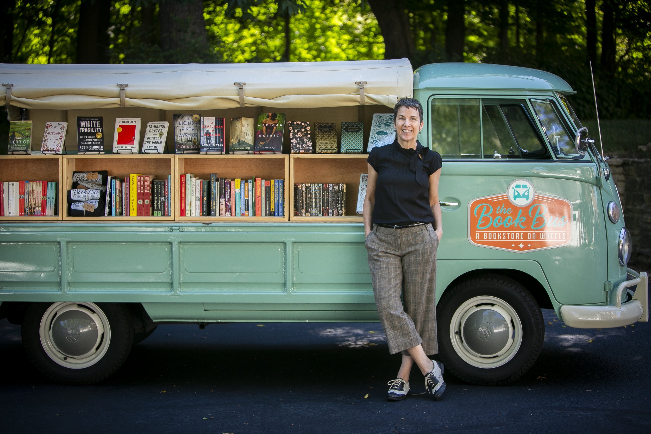 Woman standing next to a vintage green bookmobile truck with a sign that says 'the Book Bus, a bookstore on wheels' and an open side displaying shelves of books inside, in an outdoor setting with trees in the background.