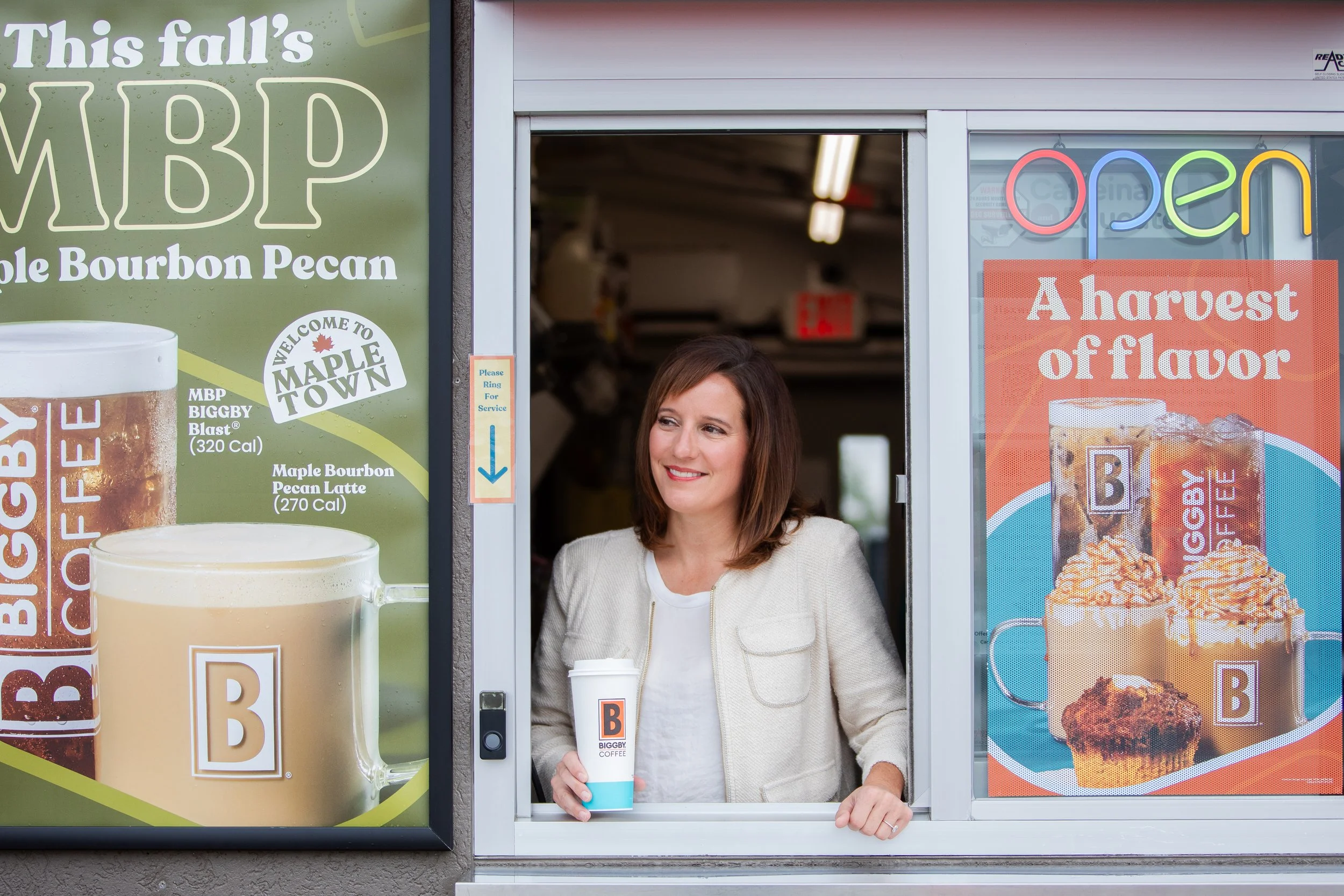 A woman standing at a drive-thru window holding a large coffee cup with a Biggby Coffee logo, smiling. The window is decorated with colorful neon 'open' sign and promotional posters for drinks and desserts.