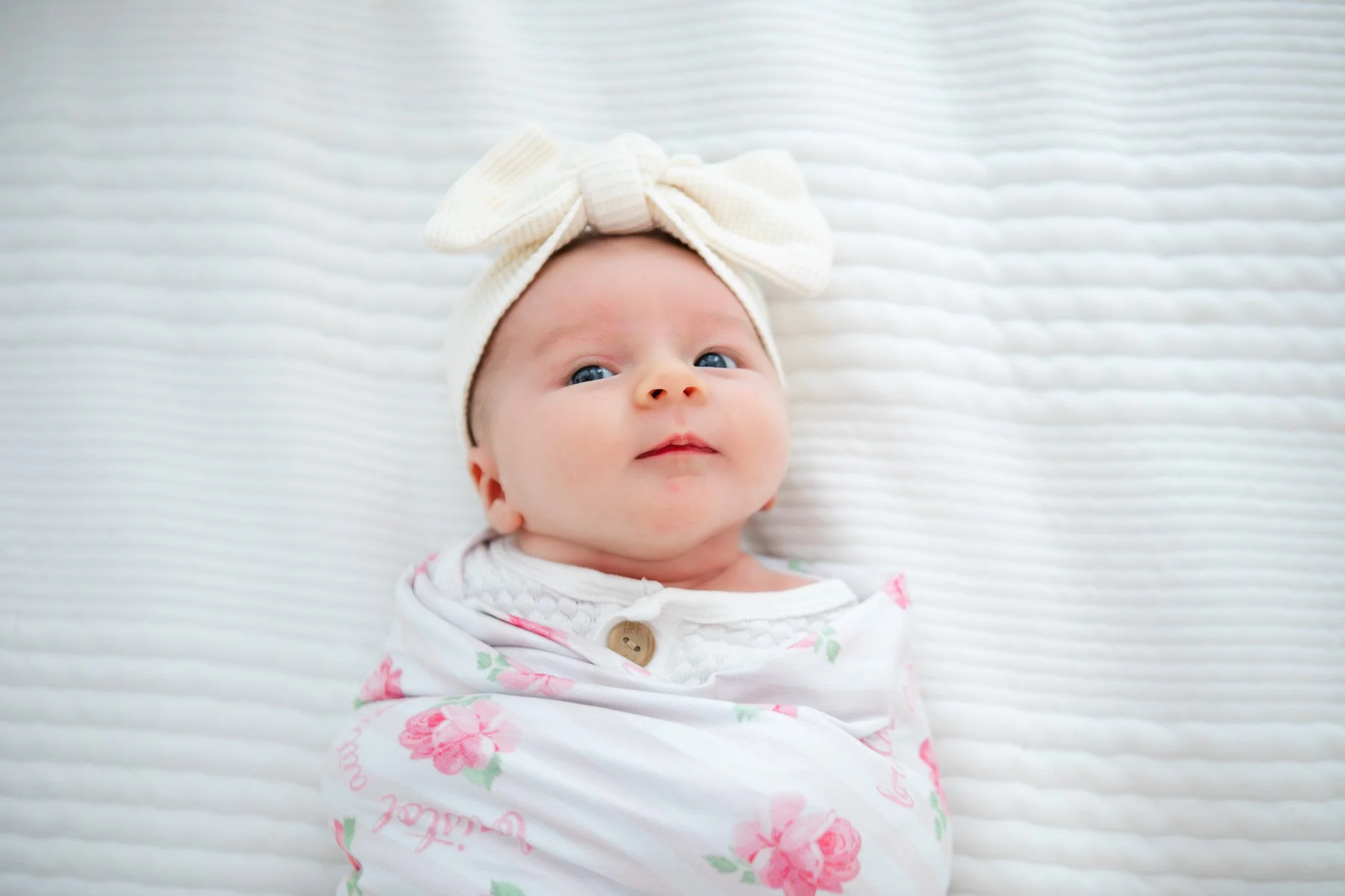 A baby wrapped in a floral blanket with a cream-colored headband and large bow, lying on a white textured surface.