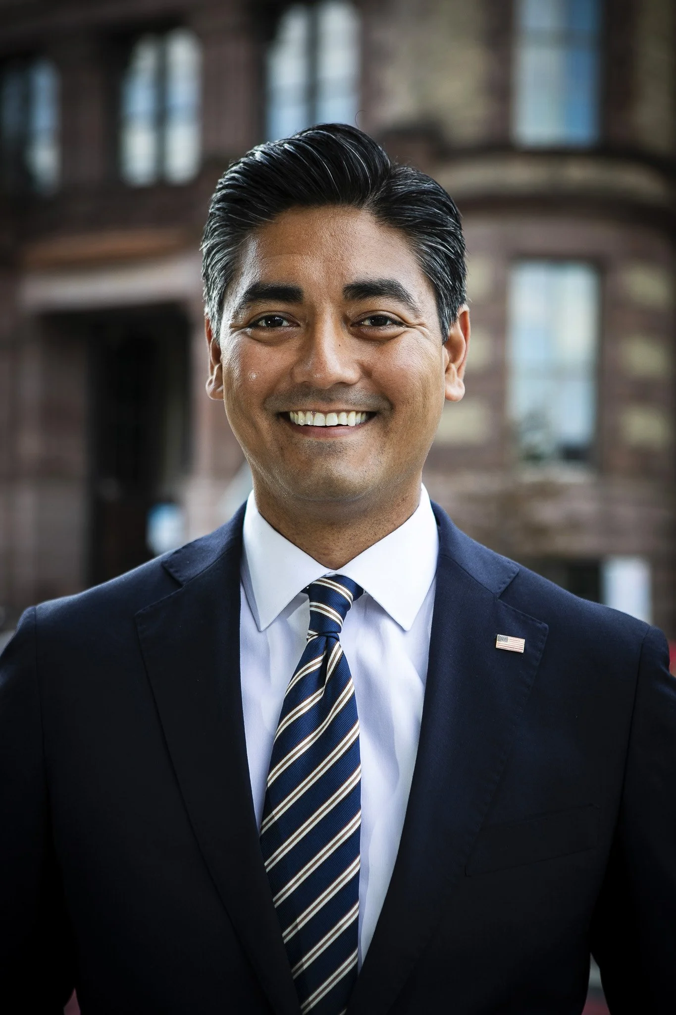 A smiling man in a business suit standing in front of a brick building with large windows.