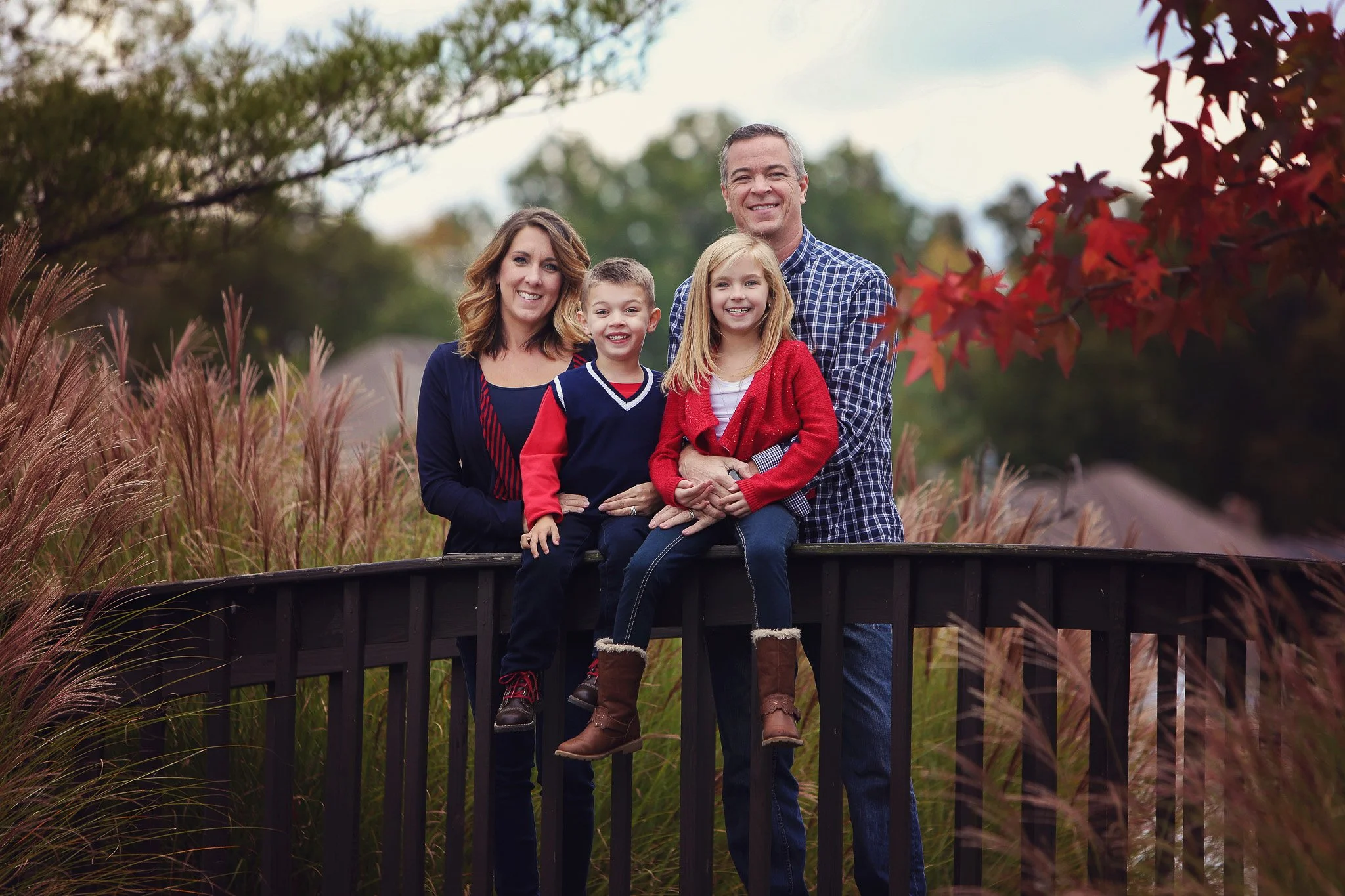 Family of four outdoors on a bridge during autumn, with trees and colorful leaves in the background.