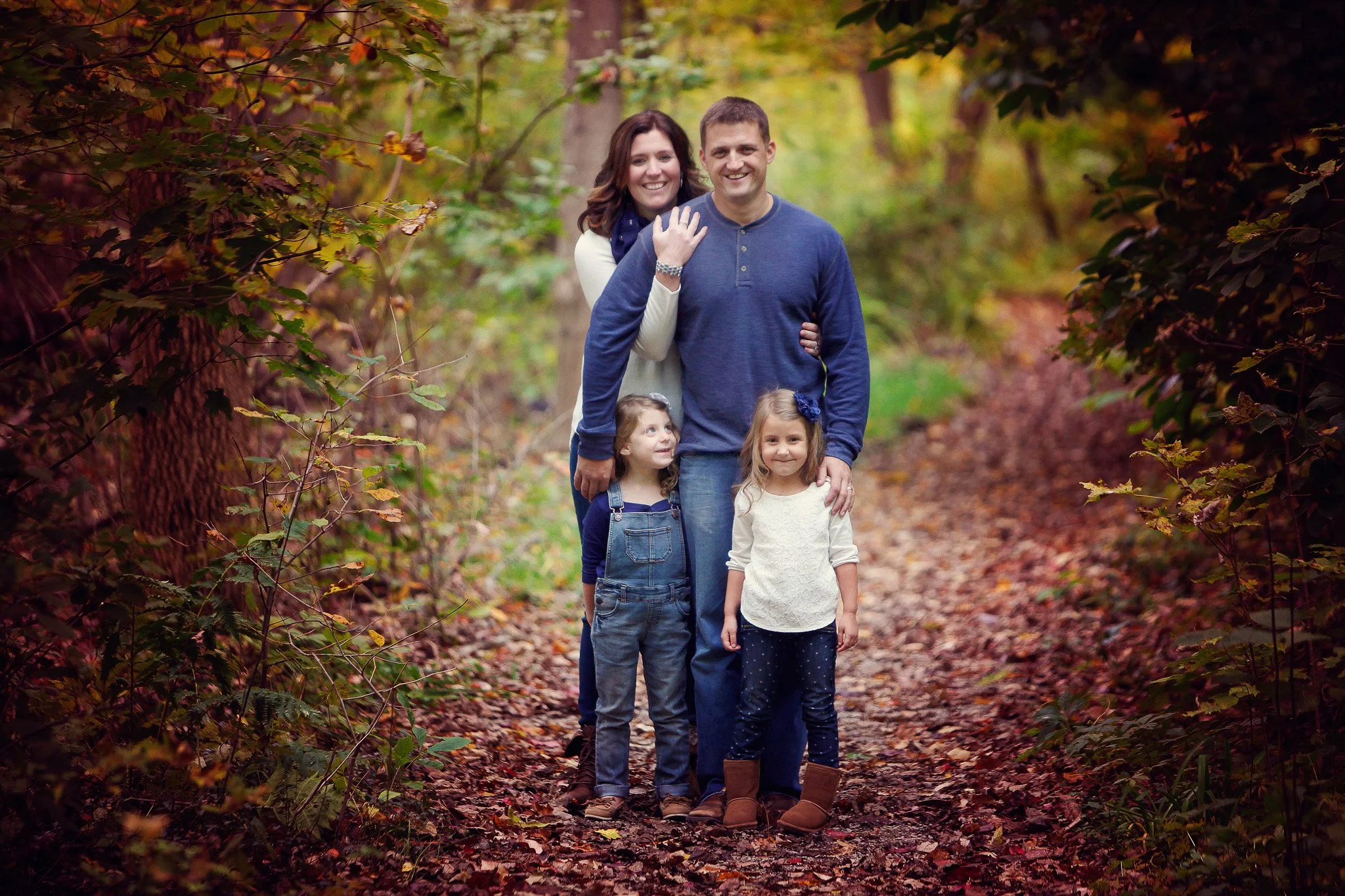 Family of four, a mother, father, and two young girls, standing on a leaf-covered forest path during autumn with colorful fall foliage in the background.