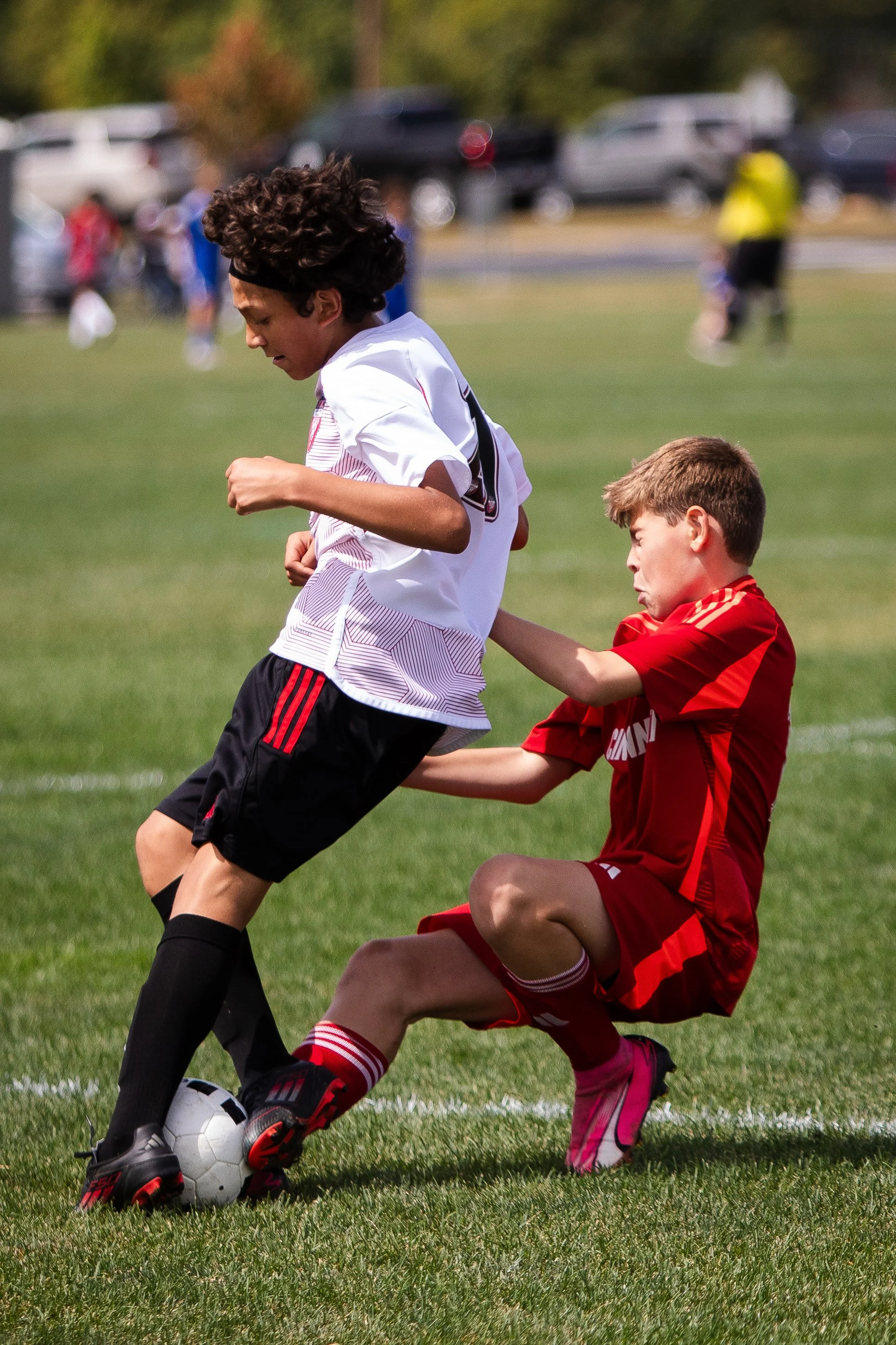 Two boys playing soccer outdoors, one in a white jersey and the other in a red jersey, with the boy in the red jersey making a sliding tackle while the boy in white controls the ball.
