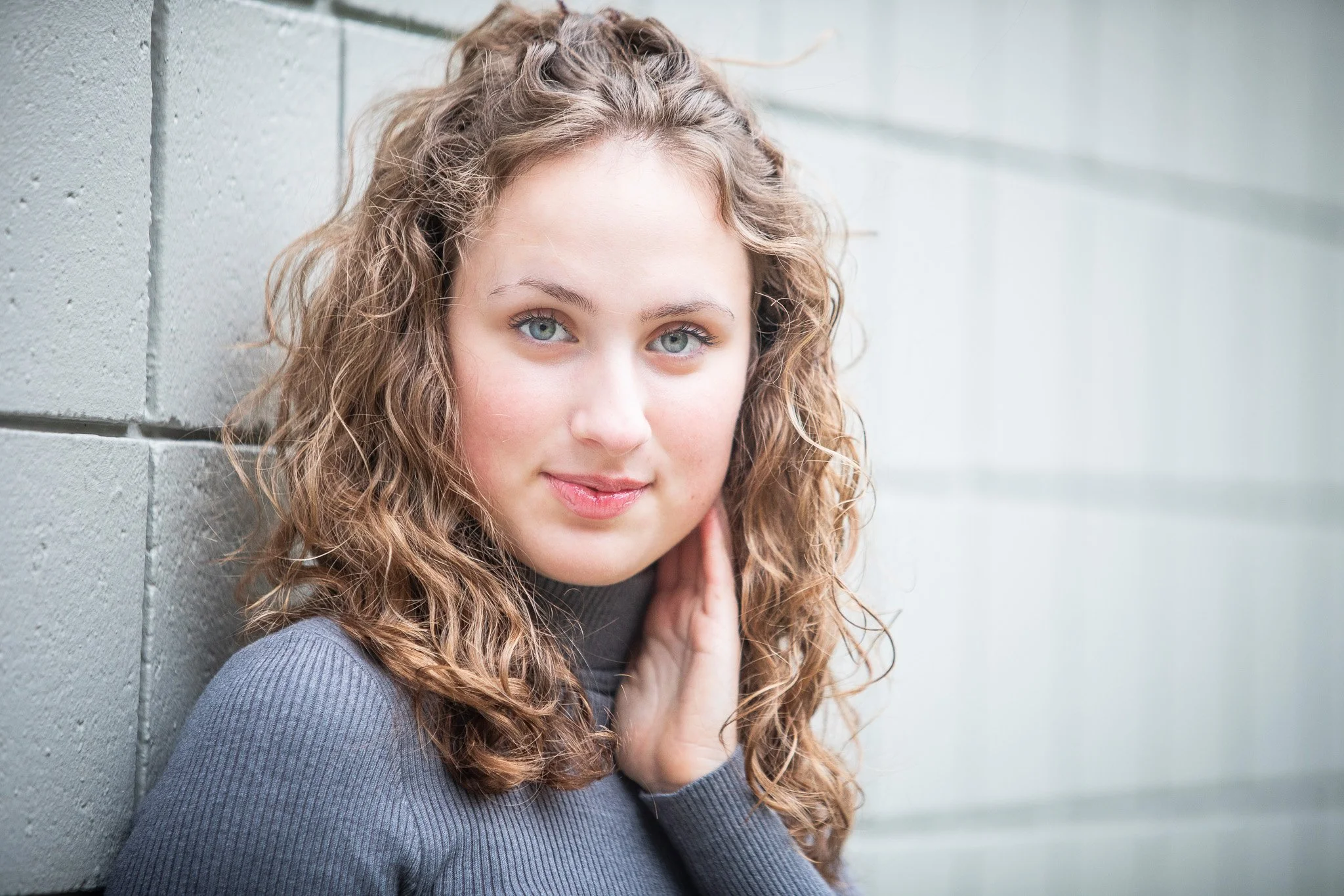 A young woman with wavy, light brown hair and blue eyes, wearing a gray top, leaning against a light green wall, looking at the camera.