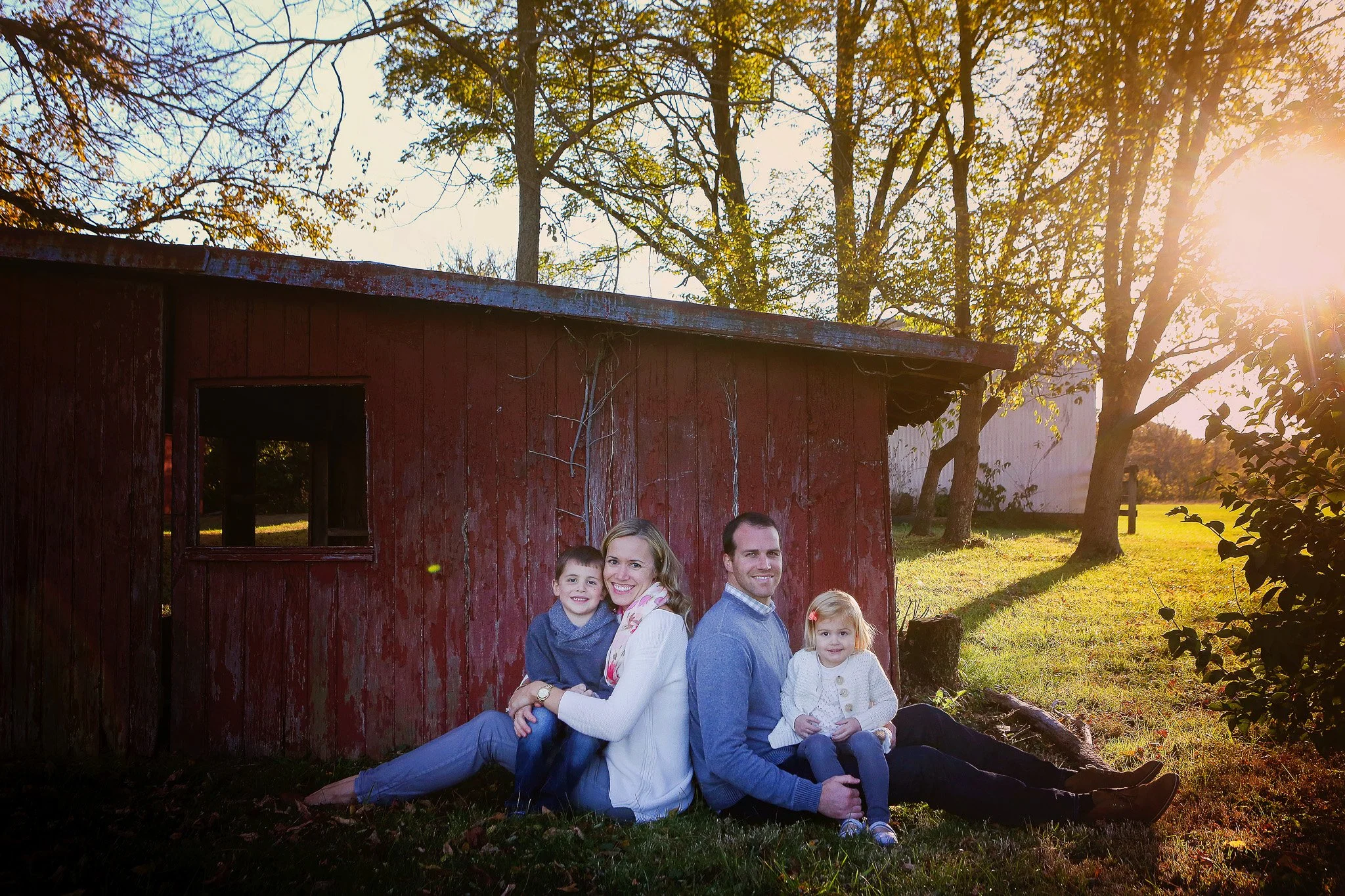 A family of four sitting on the grass in front of a weathered red shed at sunset, surrounded by trees with autumn foliage.