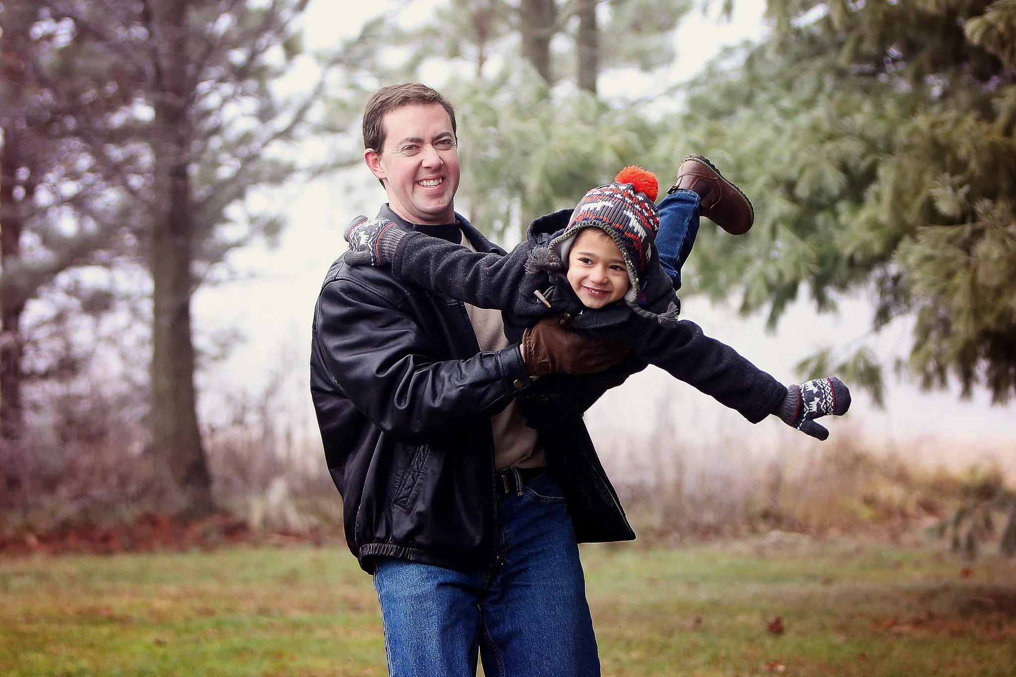 A man is holding a young boy horizontally outside in a park or wooded area, smiling and playing. The boy is dressed warmly with a colorful knit hat, gloves, and winter clothing, while the man is wearing a black leather jacket and jeans.