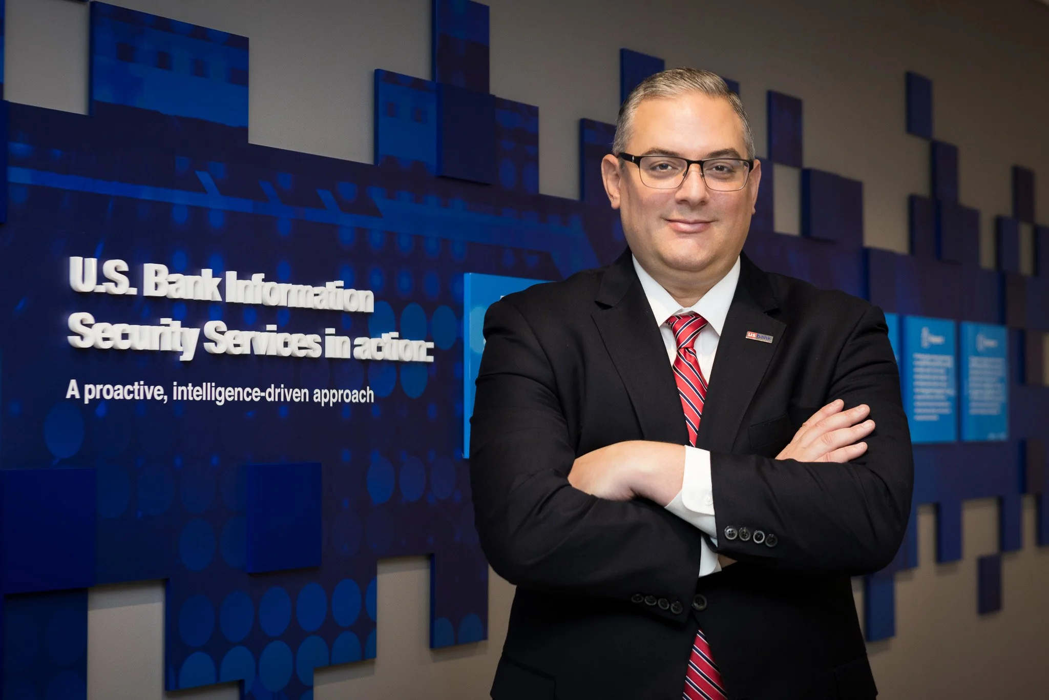 A man in a suit with glasses, standing with arms crossed in front of a sign that reads "U.S. Bank Information Security Services in action" at a corporate office.