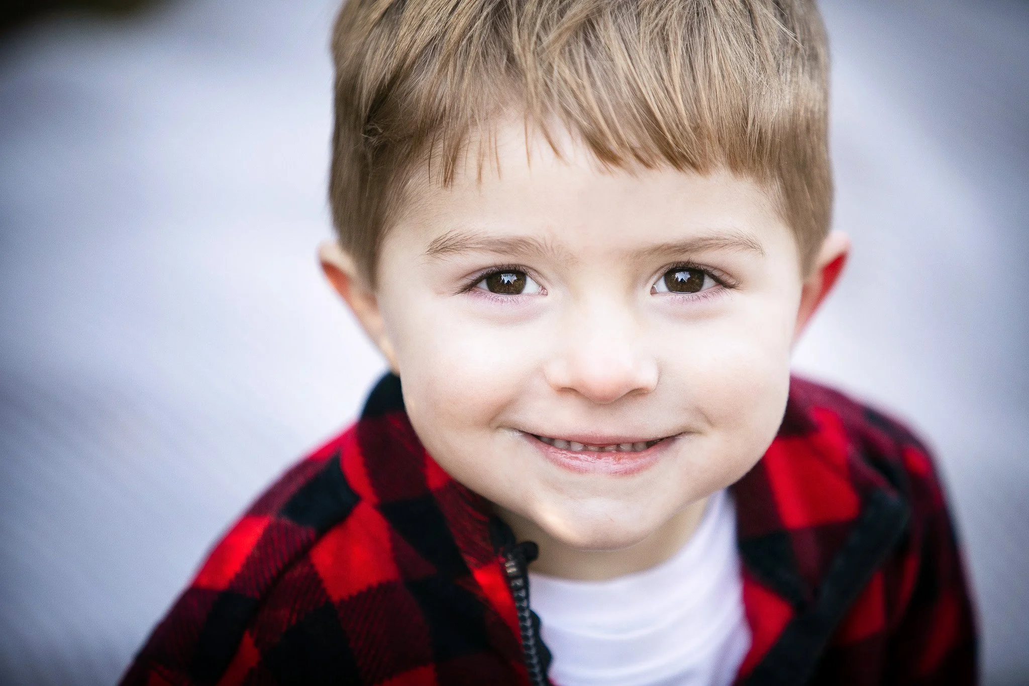 A young boy with light brown hair and brown eyes smiling at the camera, wearing a red and black checkered jacket.