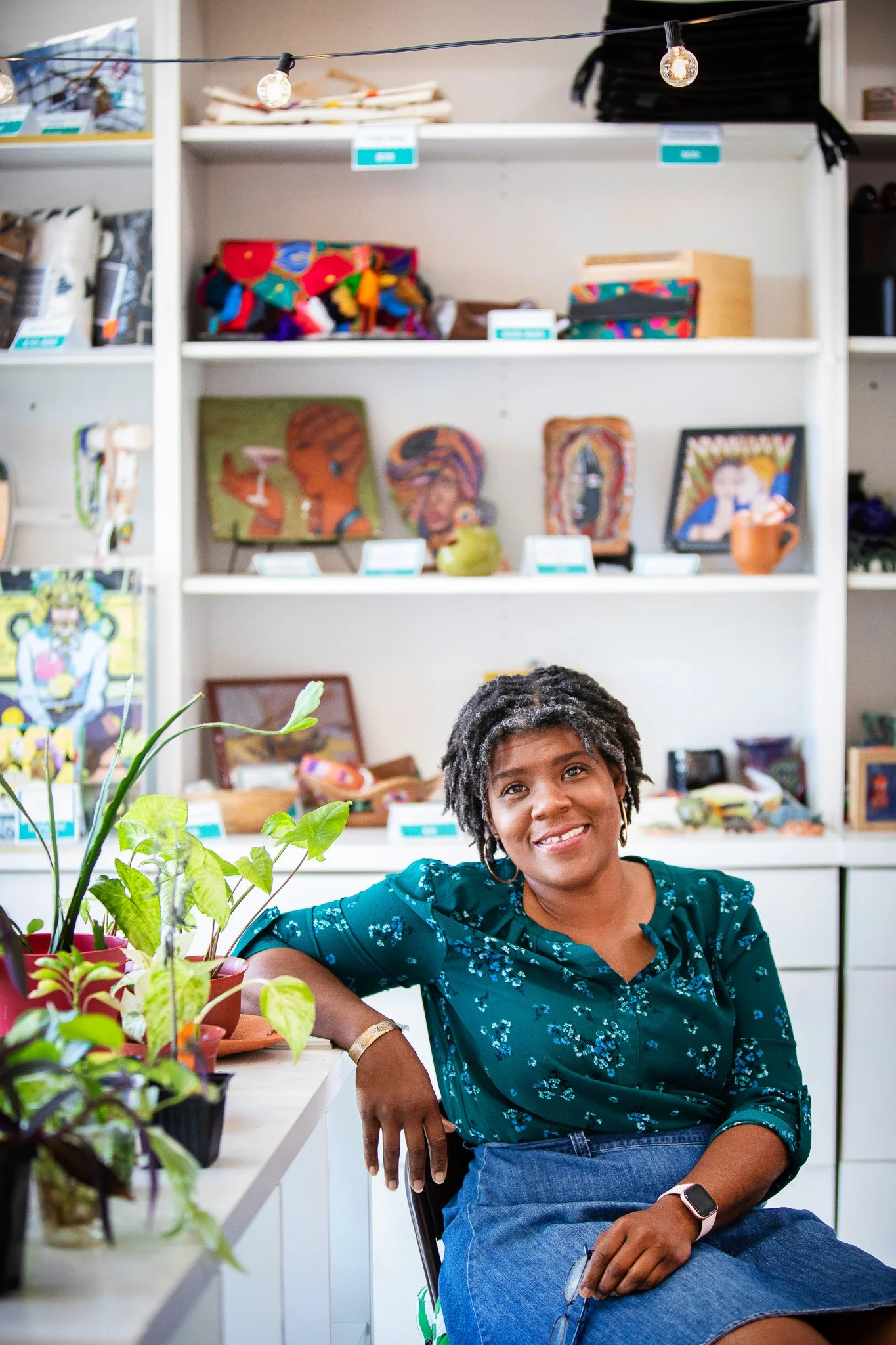 A woman with short curly hair sitting at a table with potted plants, smiling in front of a white bookshelf filled with colorful artwork, crafts, and decorations.