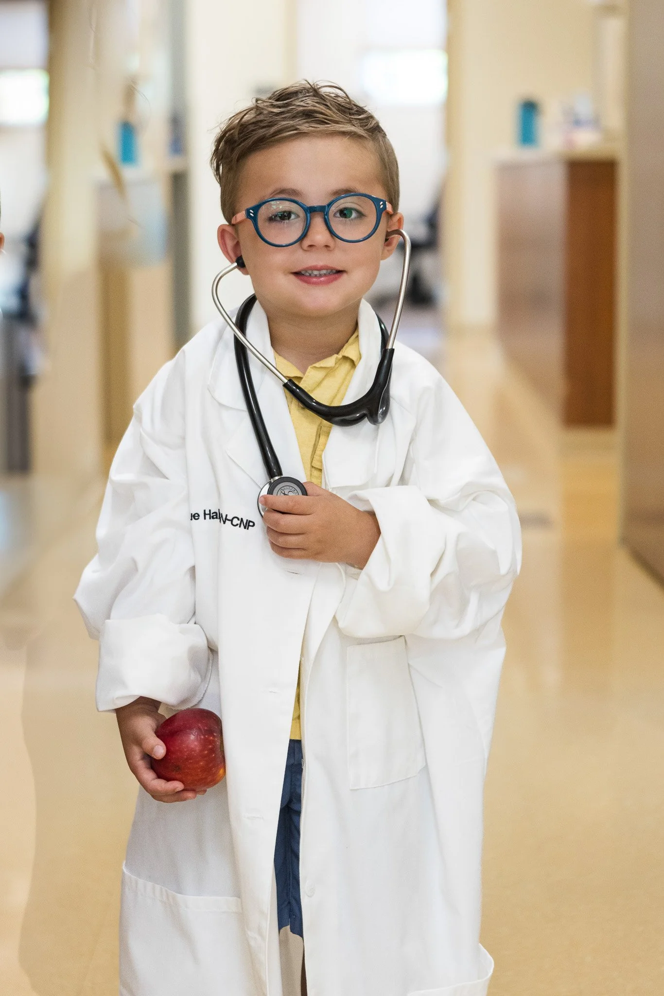 A young boy dressed as a doctor in a white coat, holding an apple, with a stethoscope around his neck, standing in a hallway.