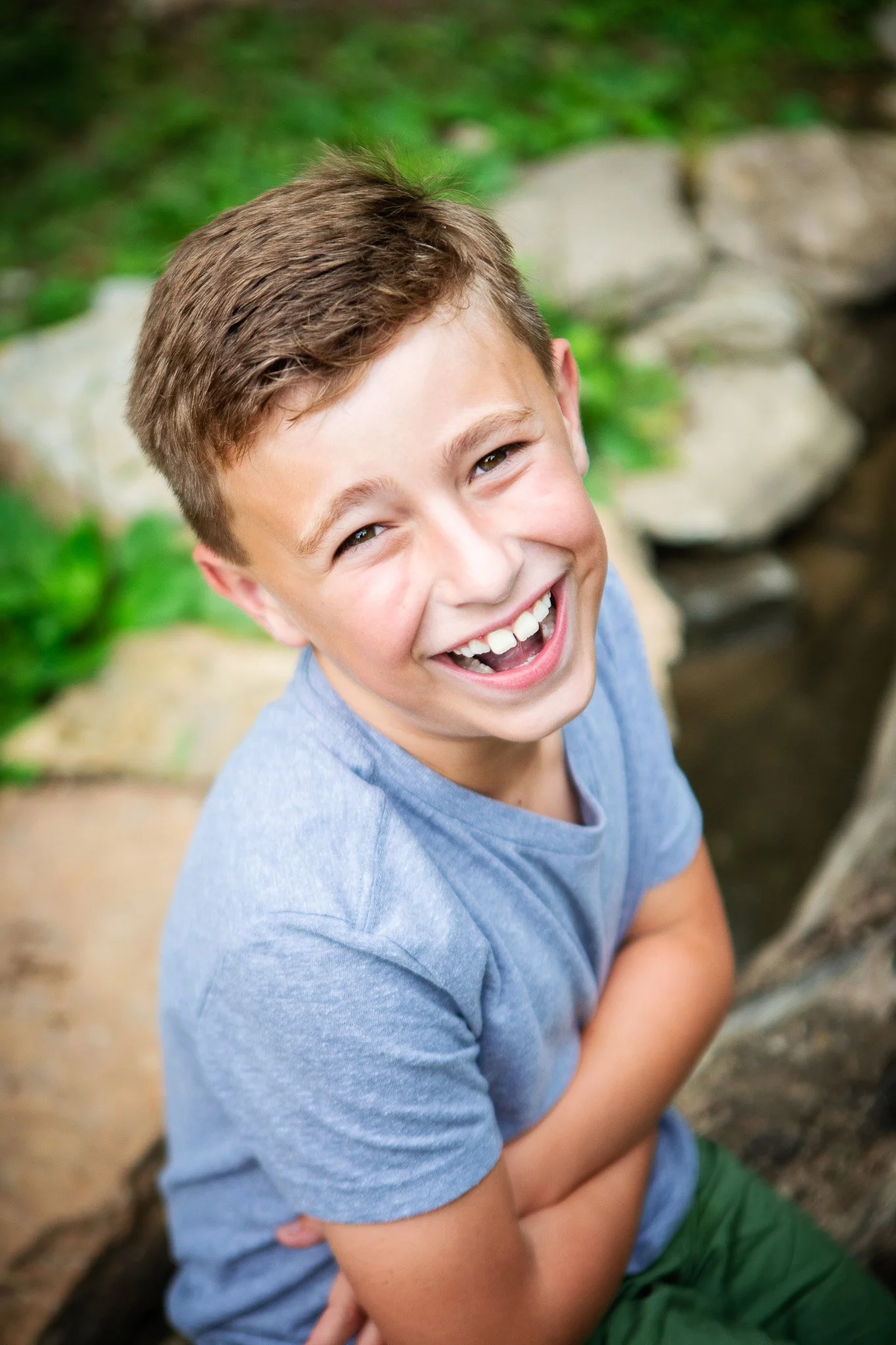 A smiling boy with short light brown hair wearing a gray t-shirt and green shorts, outdoors near rocks and greenery.