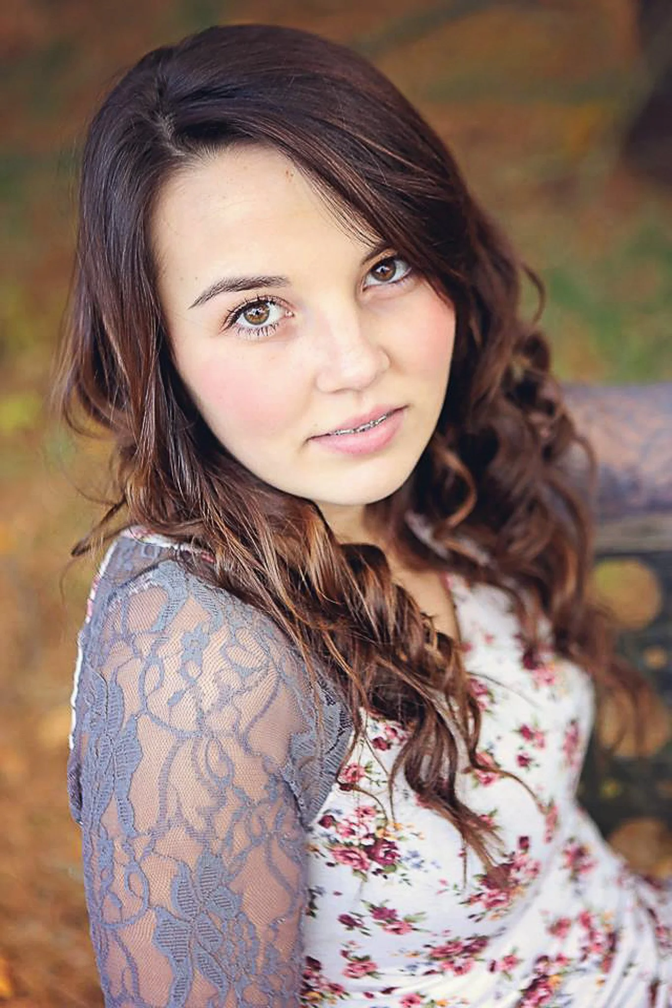 Close-up of a young woman with long, wavy brown hair, brown eyes, and light skin, wearing a floral dress with lace sleeves outdoors in autumn.