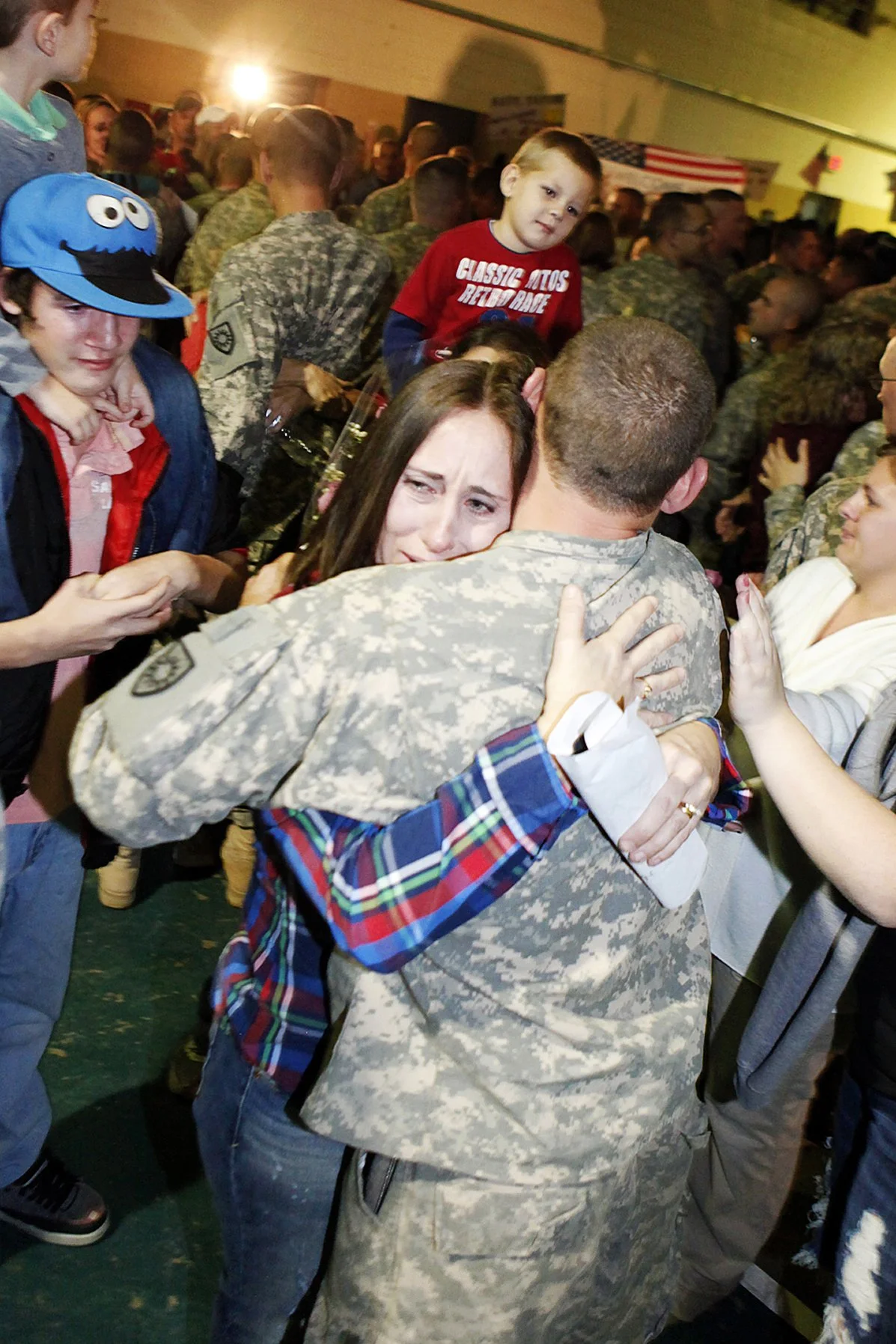 A soldier in uniform embraces a woman in a crowded indoor setting, surrounded by children and adults, some wearing camouflage and casual clothes.