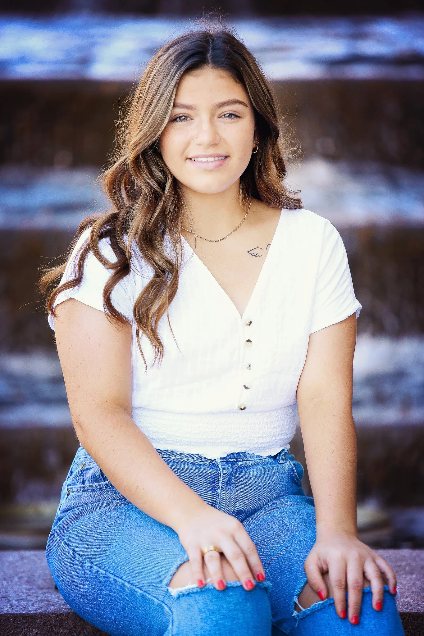Young woman with wavy brown hair, sitting outdoors near a water feature, wearing a white short-sleeved top and ripped blue jeans, smiling at the camera.