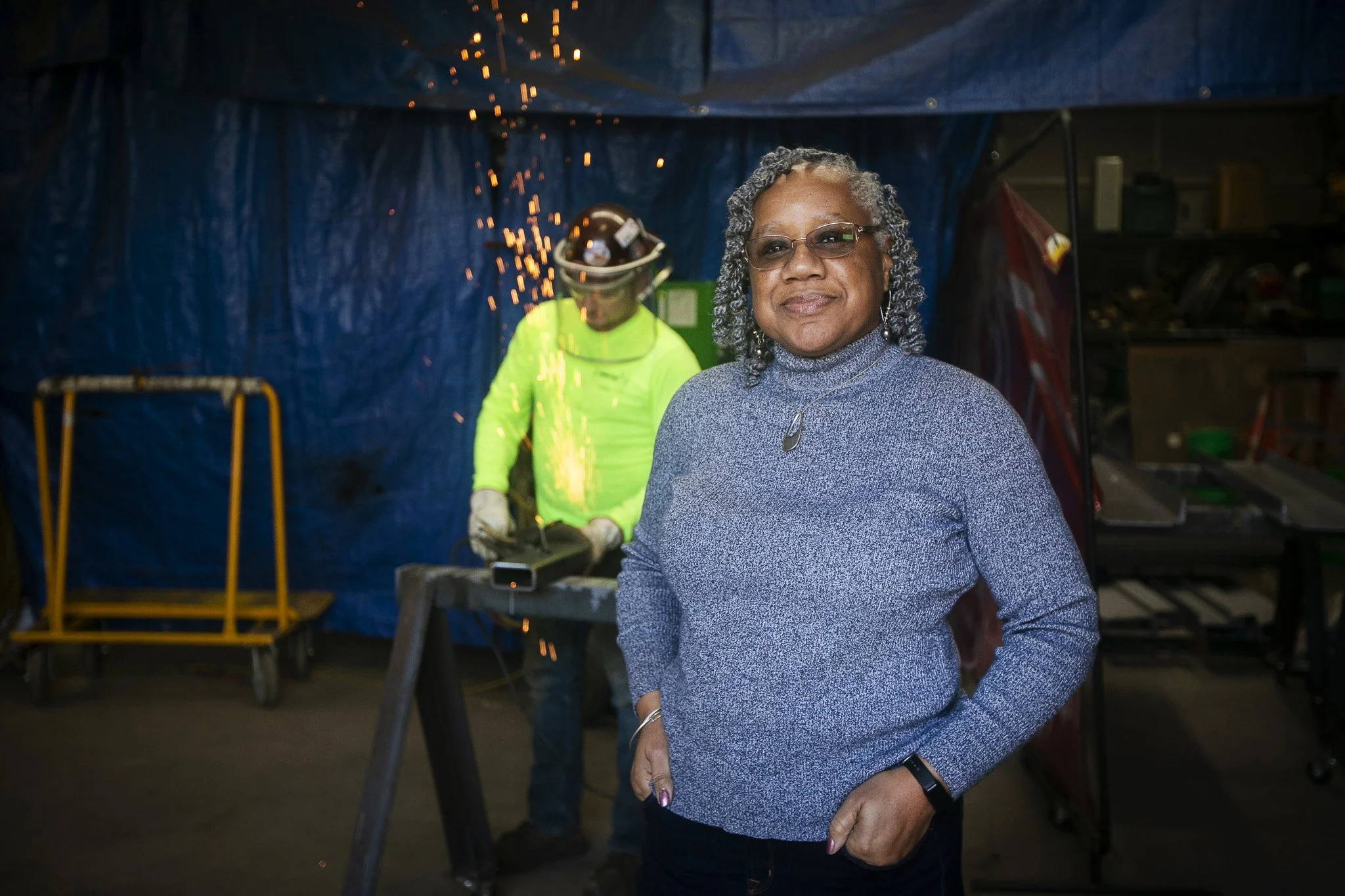 Woman with grey hair, glasses, and a grey sweater standing indoors with her hands in her pockets. In the background, a man in a bright yellow shirt and protective gear is welding, with sparks flying.