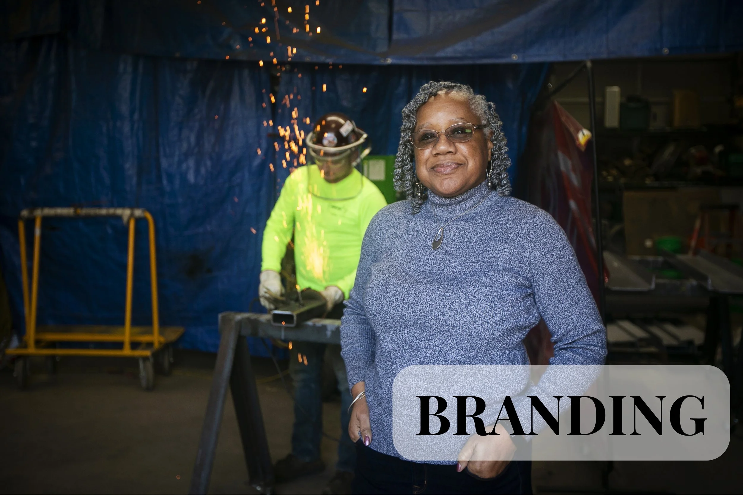 A branding portrait session in Cincinnati, Ohio. An older woman with glasses and gray curly hair standing in front of a blue tarp, with a woman welding in the background. The woman in the foreground is wearing a gray sweater and smiling slightly. The