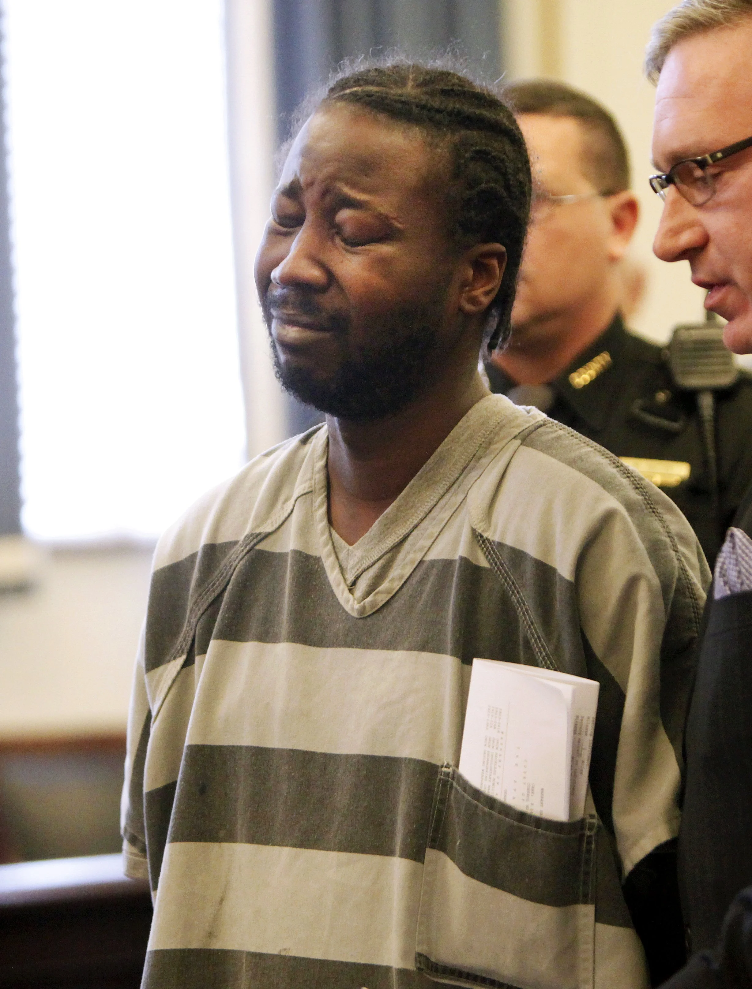 A man in a striped prison jumpsuit with his eyes closed and a distressed expression, standing in a courtroom or similar setting.