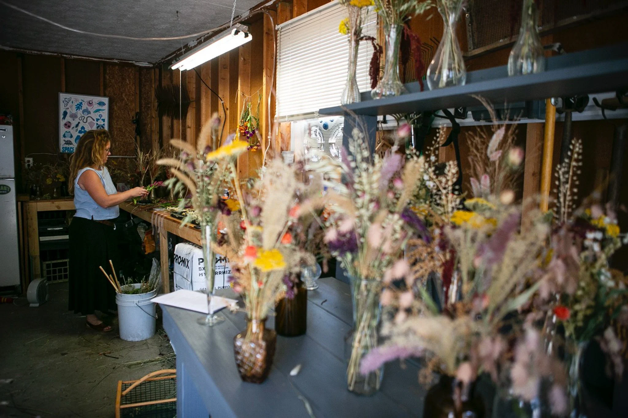 A woman arranging flowers in a workshop with wooden walls, shelves with glass vases, and a window with closed blinds.
