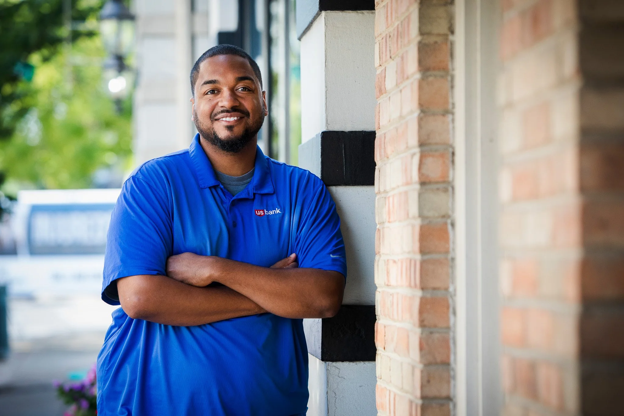 Man smiling, wearing a blue shirt with a US Bank logo, standing outdoors near a brick wall.