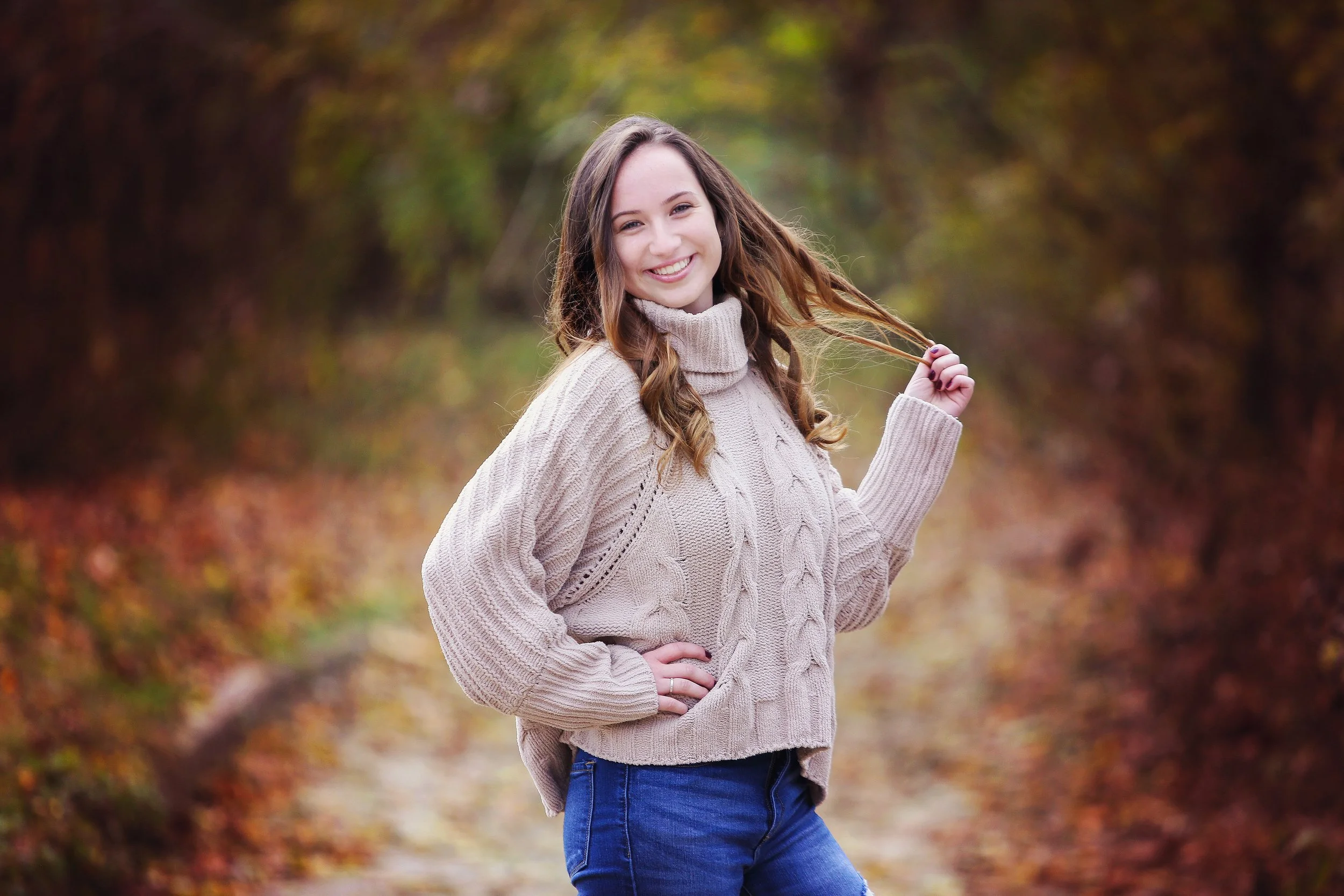 A young woman with brown hair smiling while standing outdoors on a fall day, holding a lock of her hair, wearing a beige cable-knit sweater and blue jeans, with autumn foliage in the background.