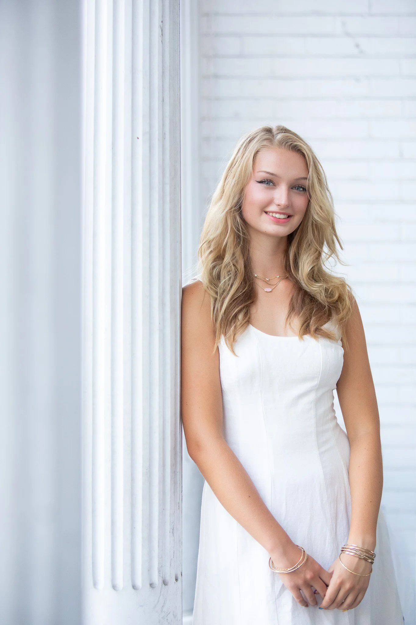 A young woman with long, wavy blonde hair, wearing a white sleeveless dress, standing near a white brick wall with a window, smiling at the camera.