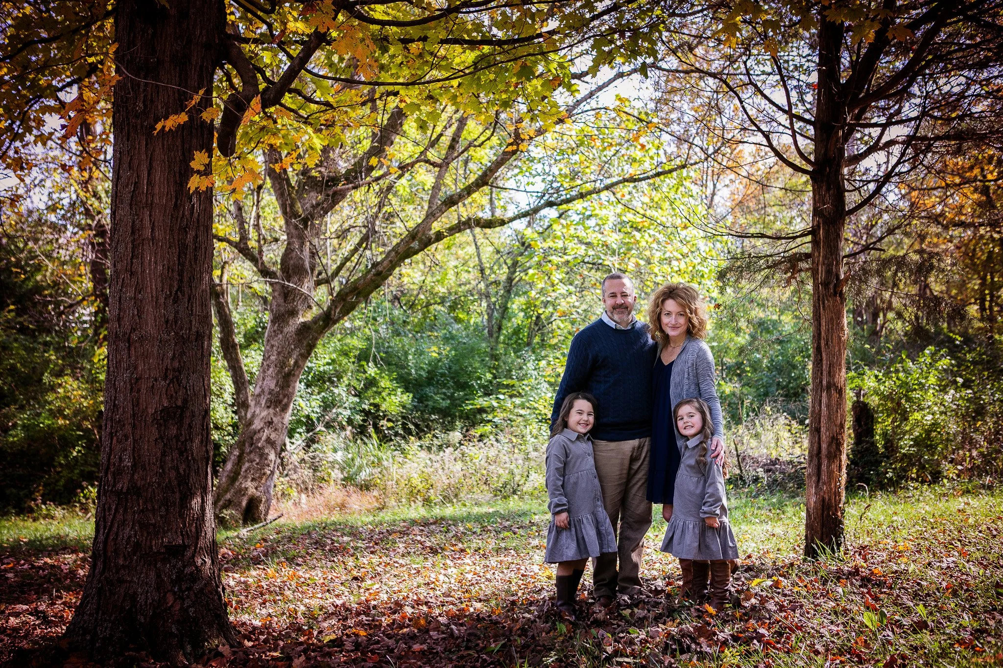 A family of four standing together outdoors in a wooded area during autumn, surrounded by trees with green and yellow leaves, and fallen leaves on the ground. The father and mother are standing behind two young girls, all smiling.