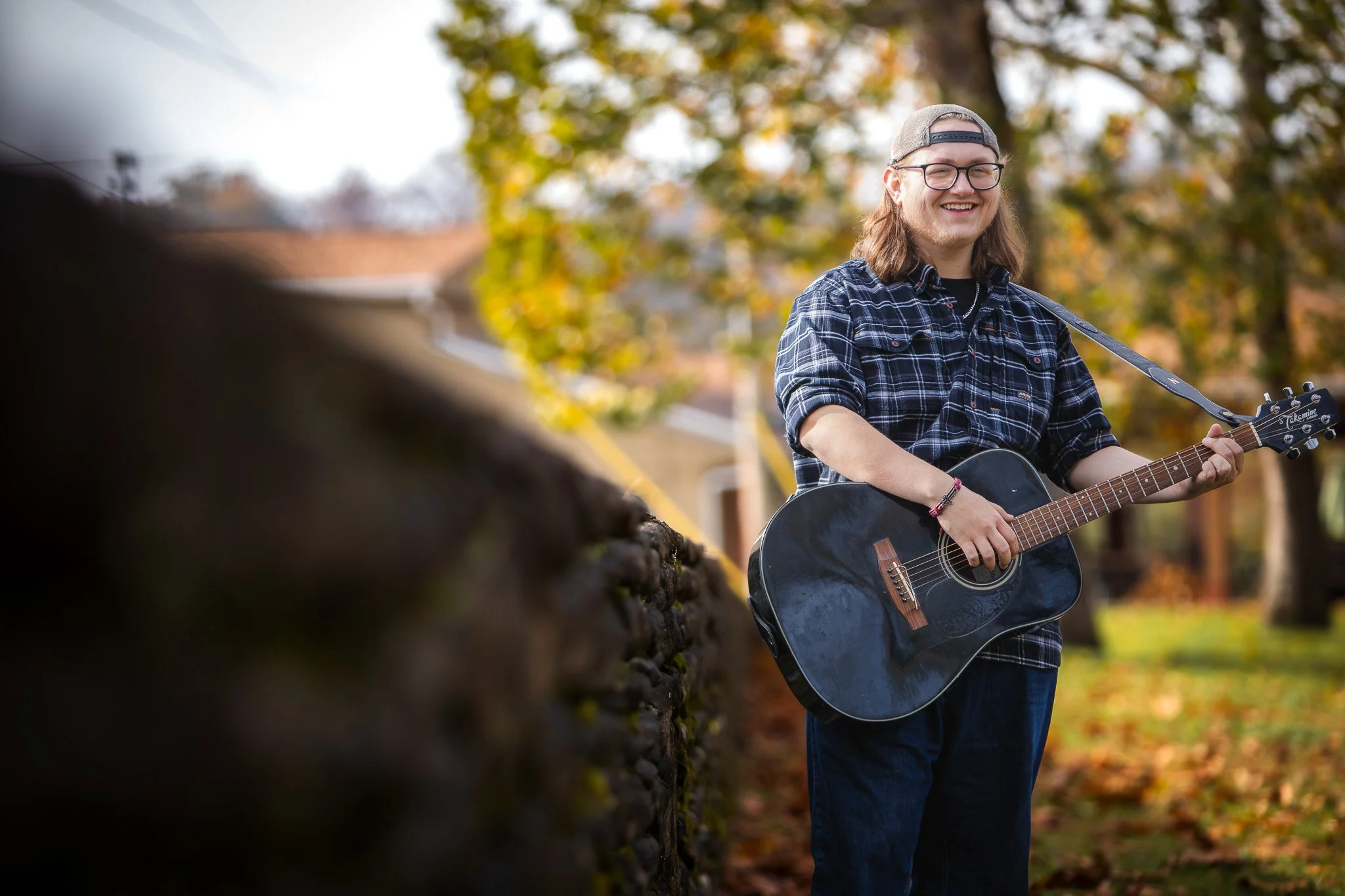 A young man with long hair, glasses, and a backwards cap, playing an acoustic guitar outdoors in a park with autumn trees.