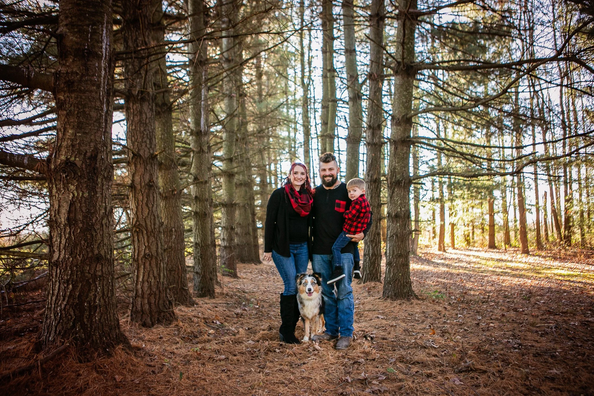 A family of three with a dog in a forest during daytime. The woman has red hair and is wearing a black coat and a red scarf. The man has a beard and is wearing a black jacket, holding a young boy dressed in a red and black checkered shirt. The dog is