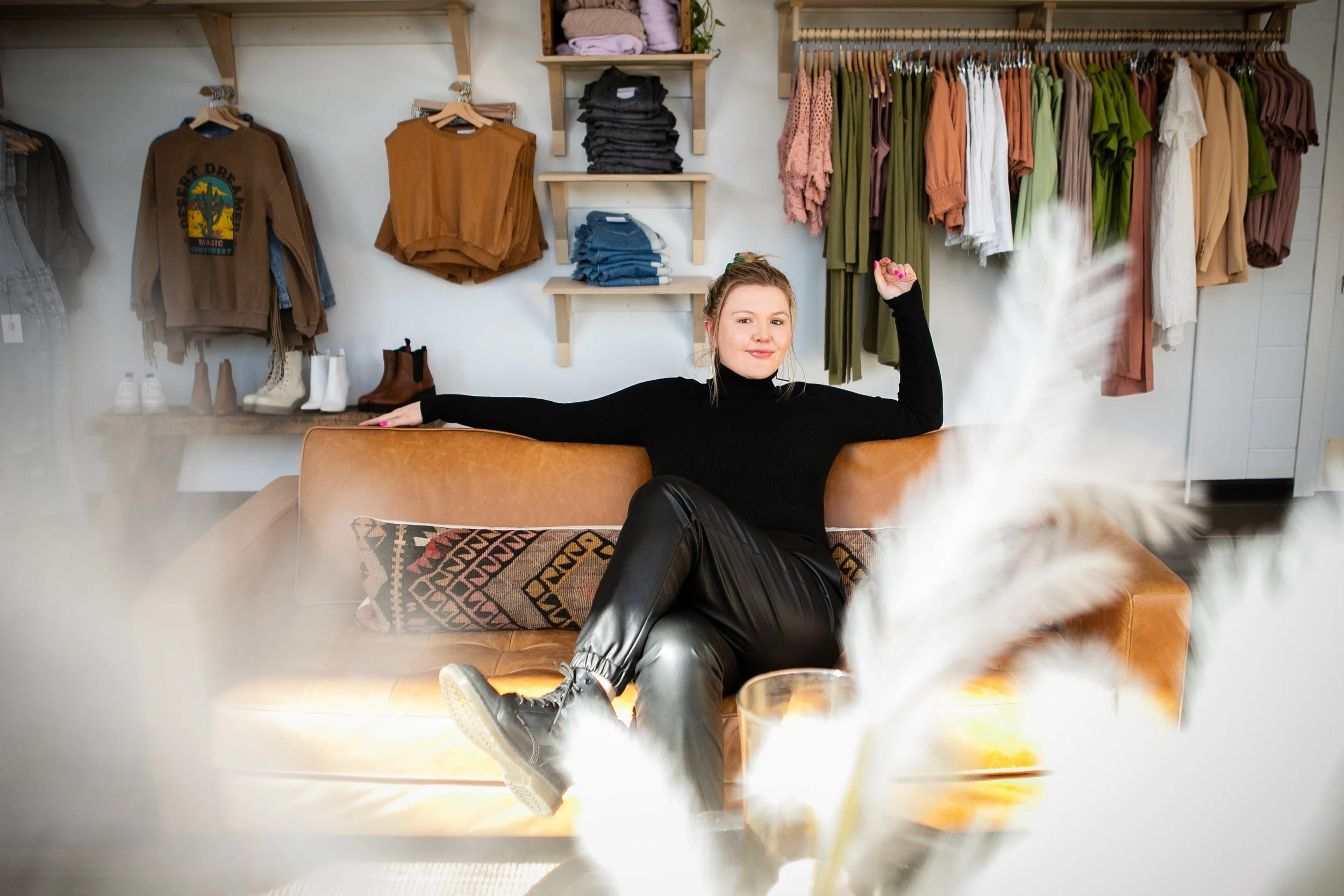 A woman sitting on a brown couch in a clothing boutique, surrounded by clothing racks and shelves, with white pampas grass in the foreground.