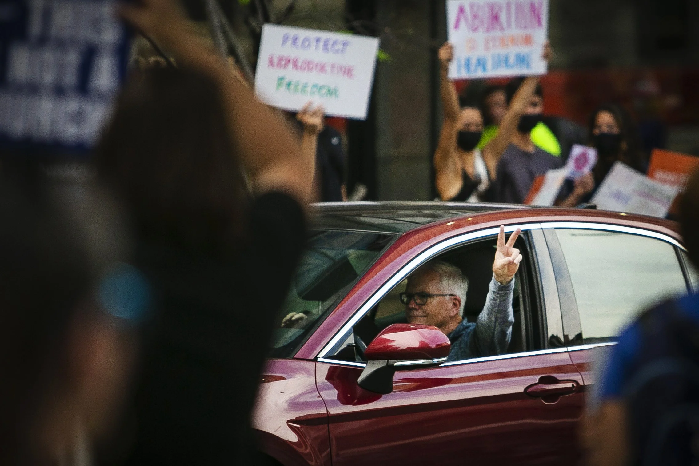 A person with glasses and gray hair inside a red car raising two fingers in a peace sign during a protest or demonstration, with people holding signs in the background.