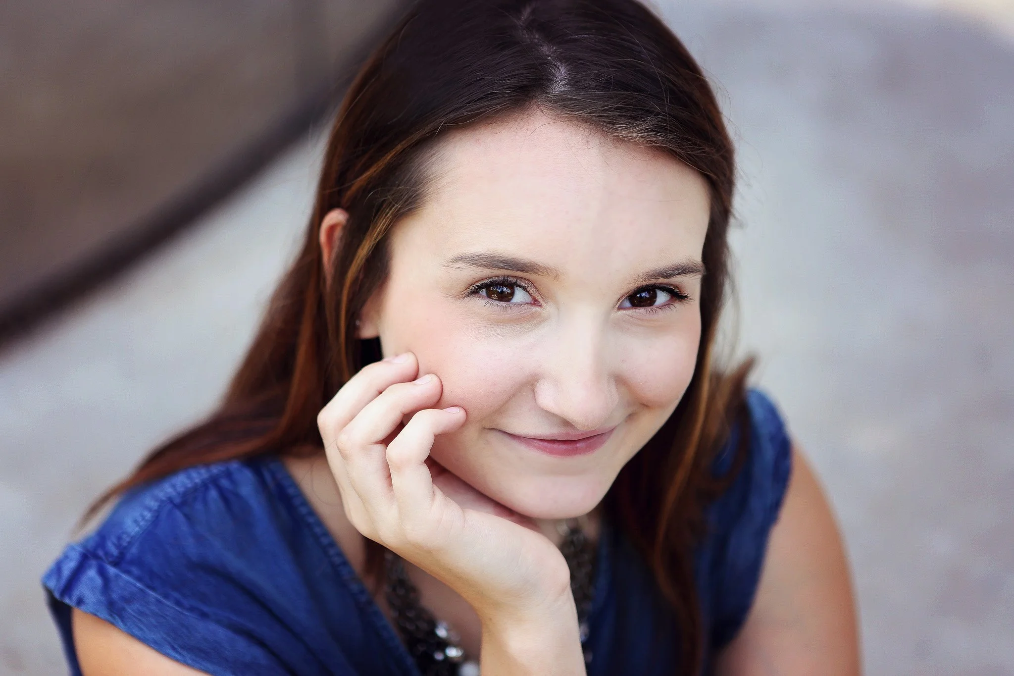 A young woman with brown hair, smiling gently, wearing a blue top, resting her chin on her hand.